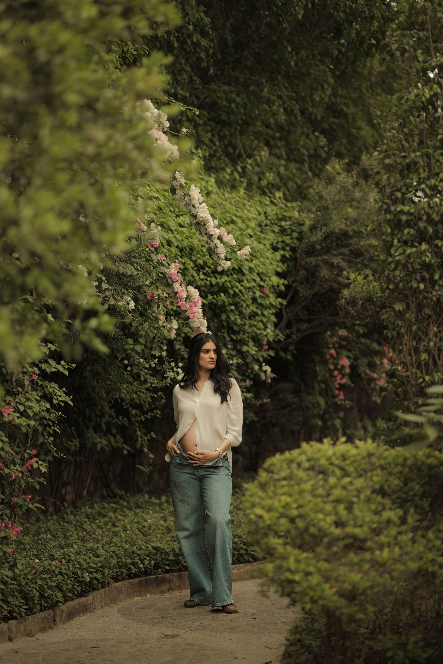 Pregnant woman in white top and jeans standing on garden path surrounded by lush green hedges and pink flowering shrubs.