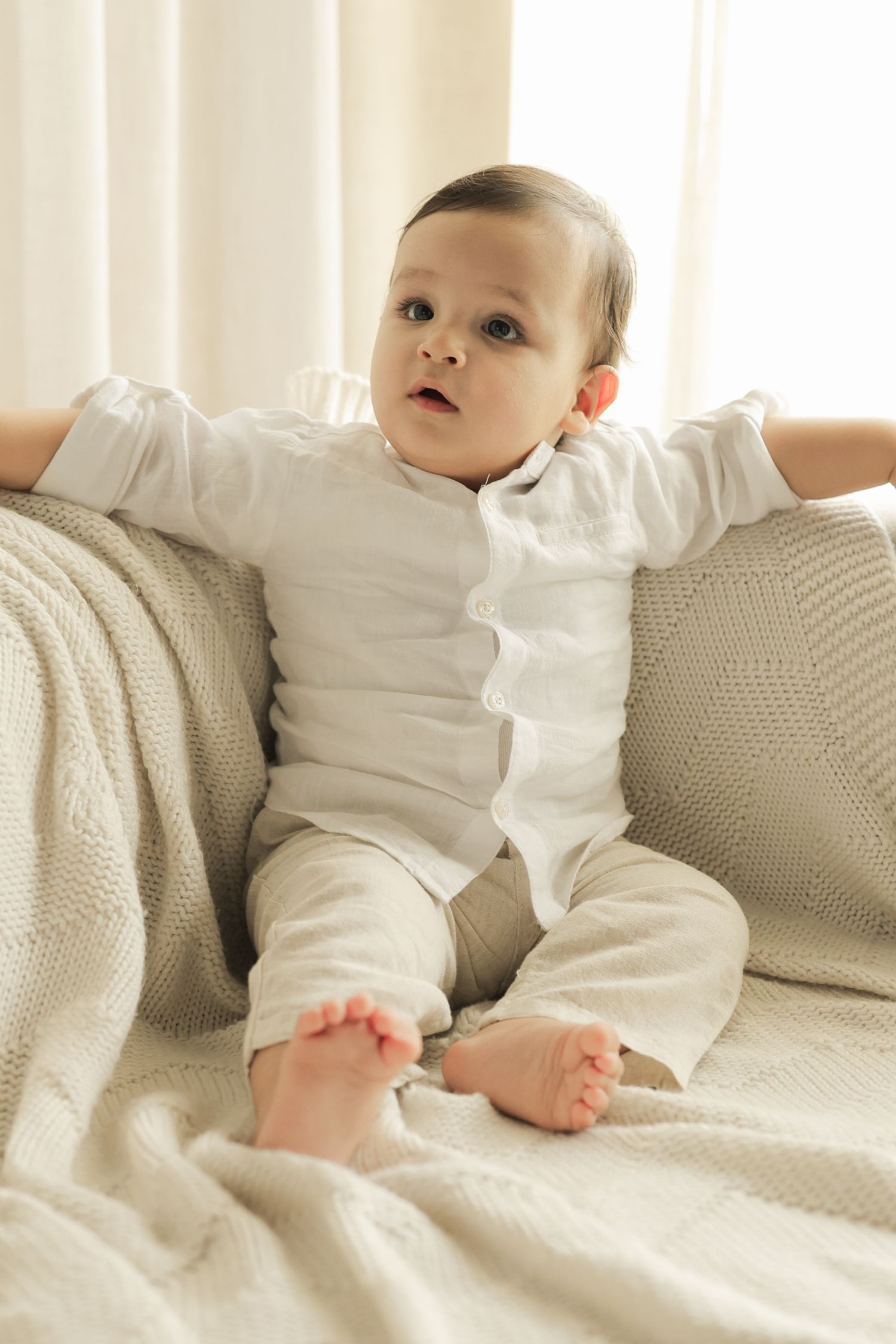 Adorable baby in white outfit sitting on a beige sofa with a knit blanket, looking curiously to the side.