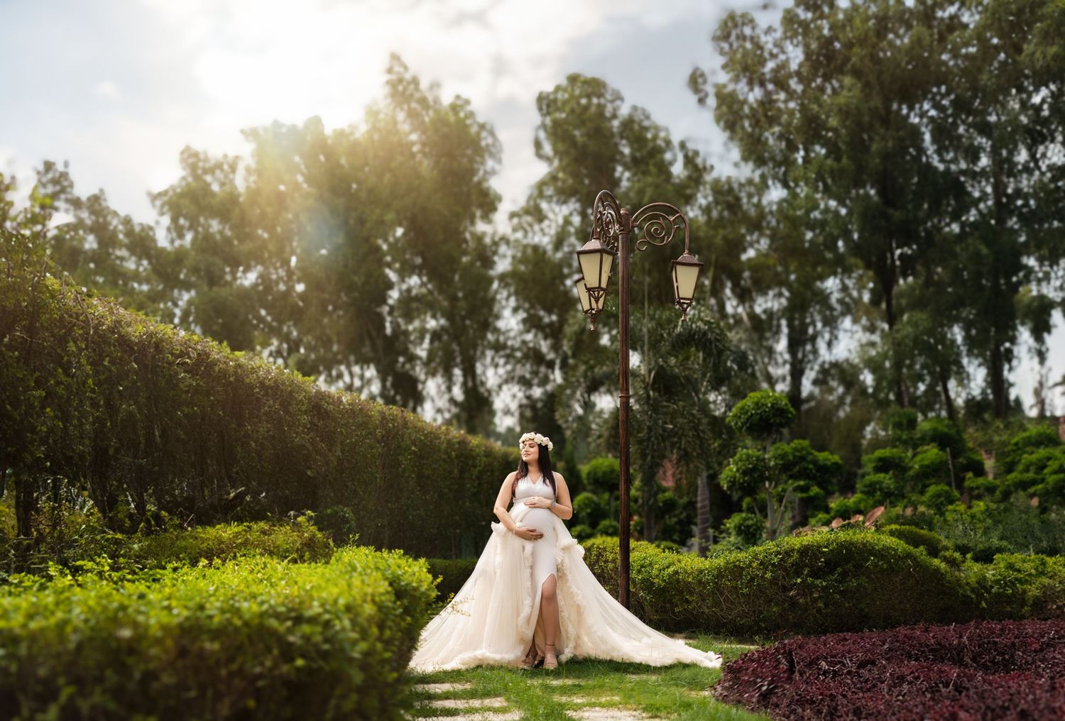 Pregnant woman in elegant pink gown posing in lush garden with ornate lamp post and sunlit trees.