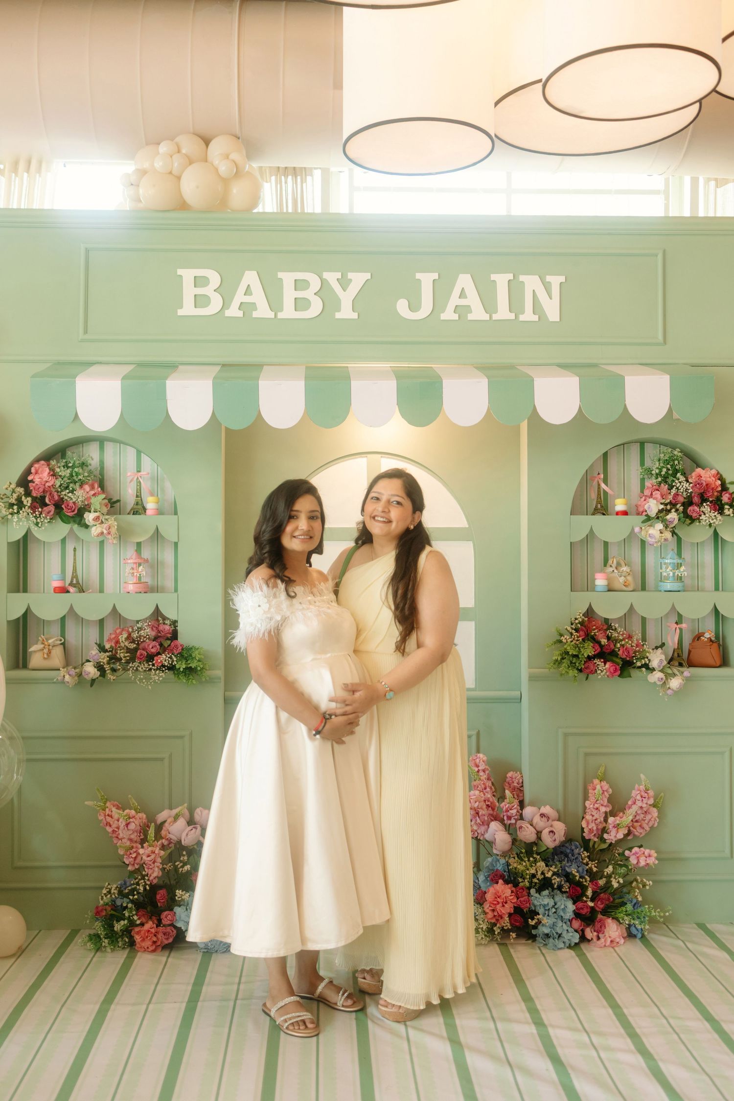 Two women pose at a Baby Jain baby shower with mint green shop backdrop, floral arrangements, and elegant cream dresses.