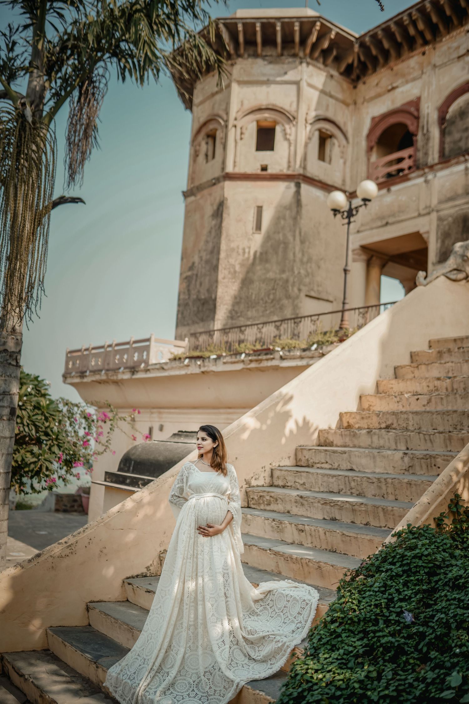 A bride in a flowing lace dress poses on historic stone steps against a Mediterranean architectural backdrop.