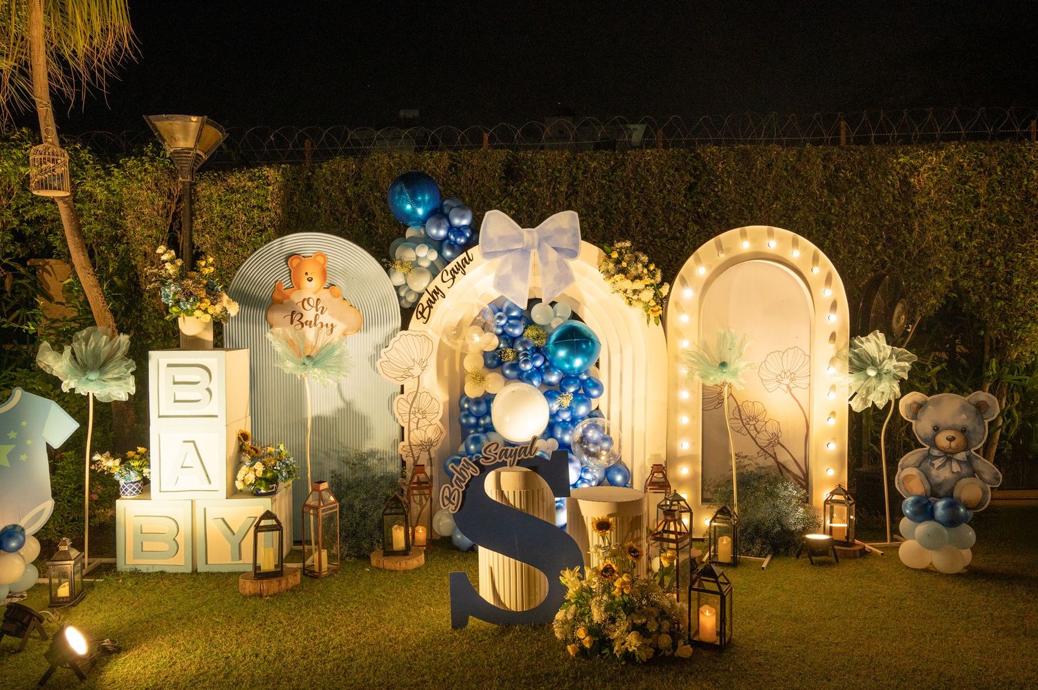 Blue and white baby shower decor with balloon arch, teddy bears, and illuminated letter blocks at night.