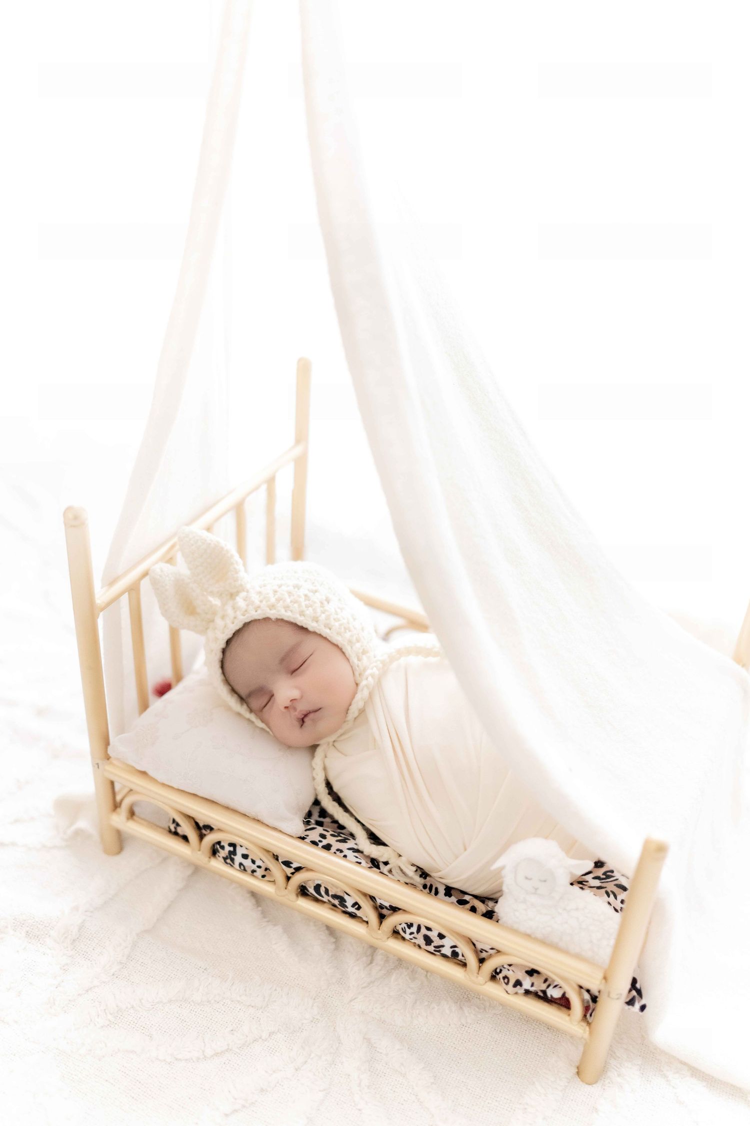 Sleeping newborn baby wrapped in white swaddle with bunny bonnet in a small wooden canopy bed.