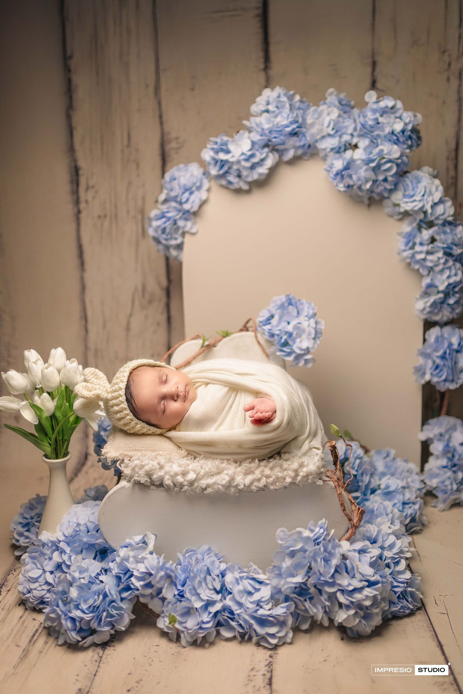 Newborn baby wrapped in white knit swaddle sleeping in a bowl surrounded by blue hydrangeas and white tulips.