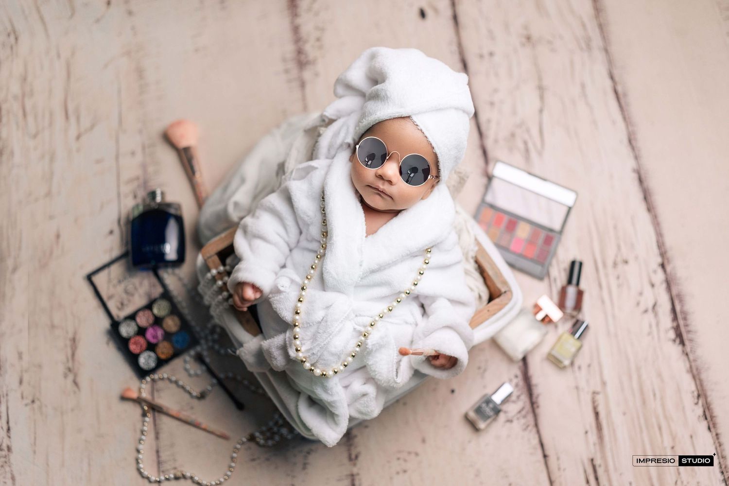Adorable newborn baby dressed in white bathrobe, towel turban, sunglasses, pearls, surrounded by makeup products.