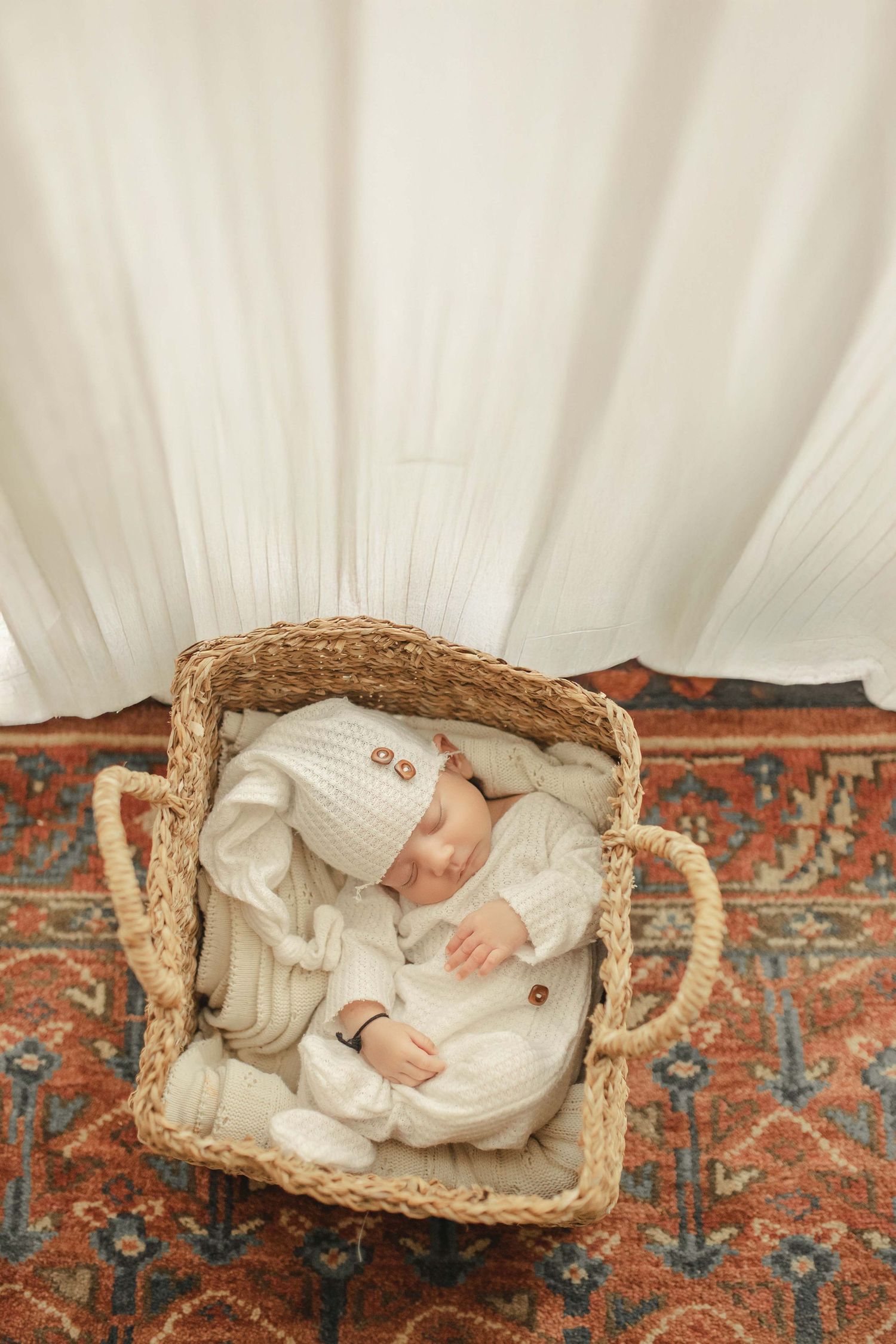 Sleeping newborn in white knit outfit and hat resting in a wicker Moses basket on a patterned rug.