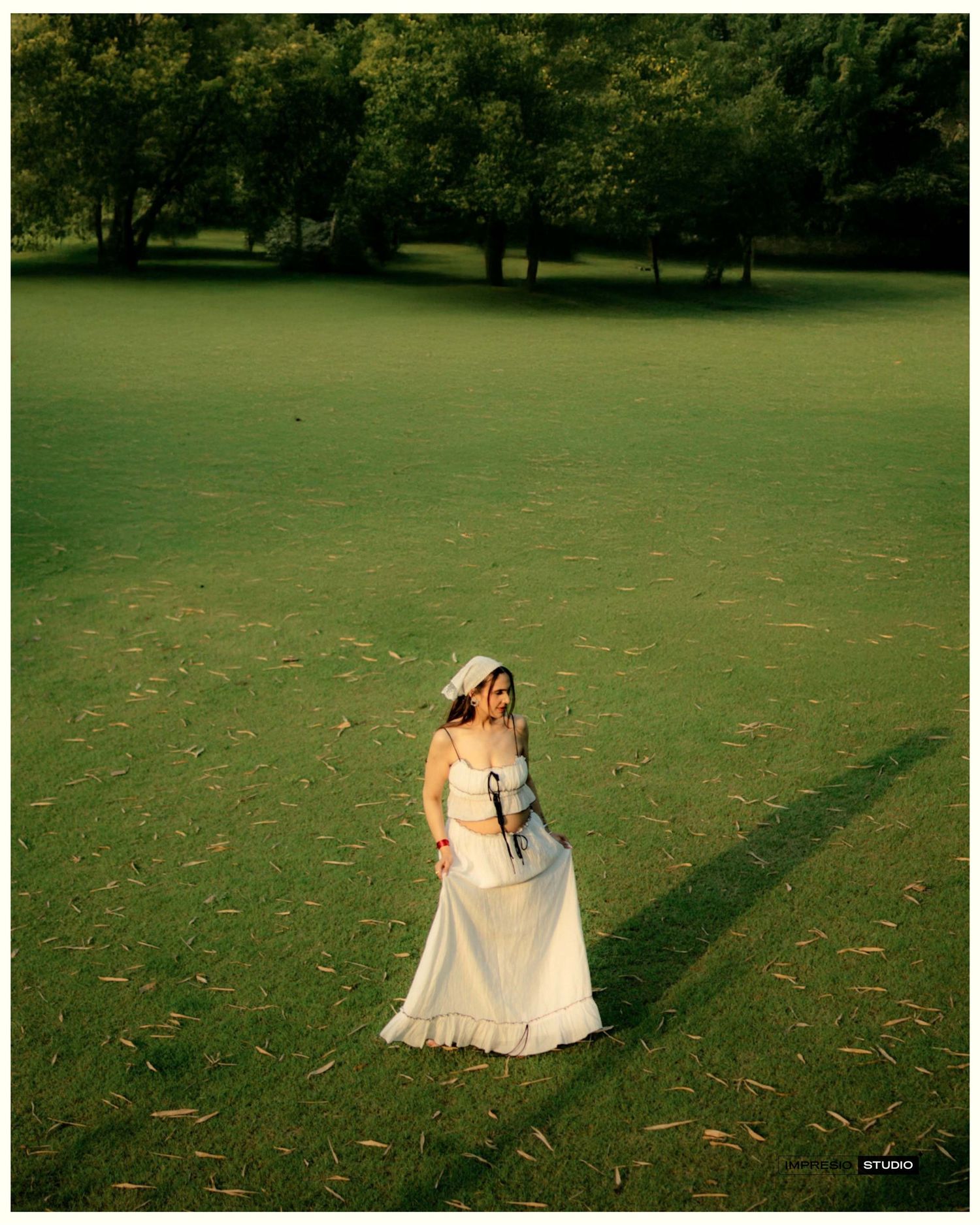 A woman in a white bridal gown and hat stands on a lush green lawn surrounded by trees.