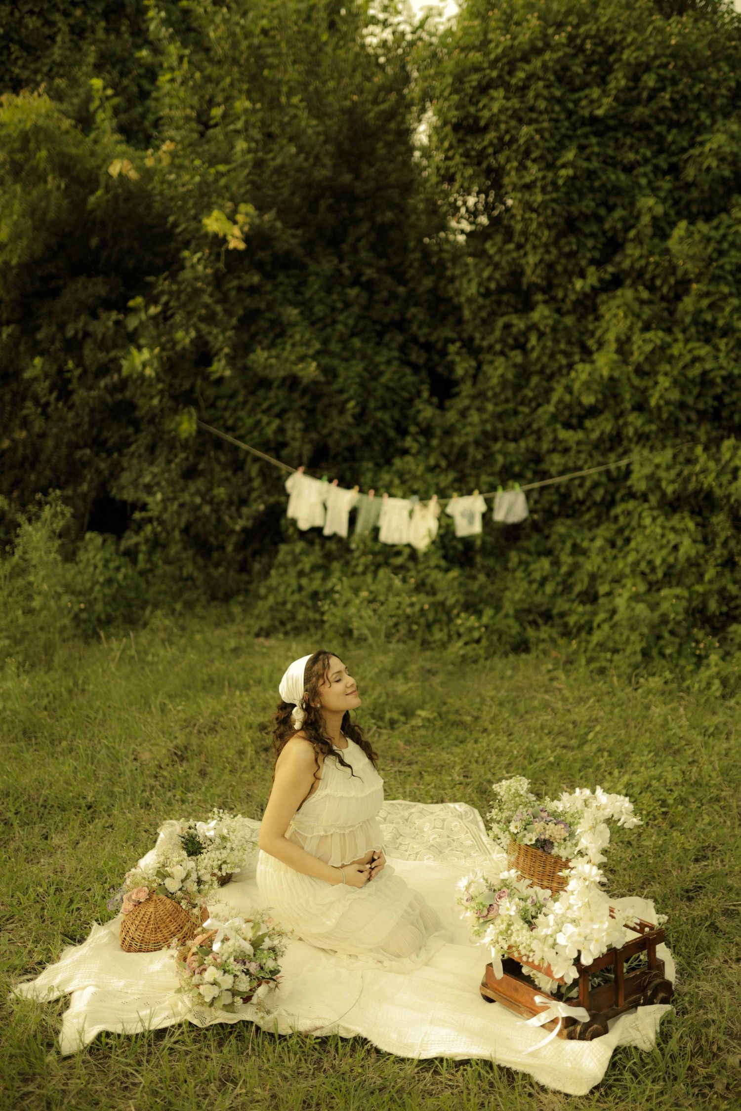 Pregnant woman in white dress sitting on blanket surrounded by flowers and baskets in a lush garden with baby clothes hanging.