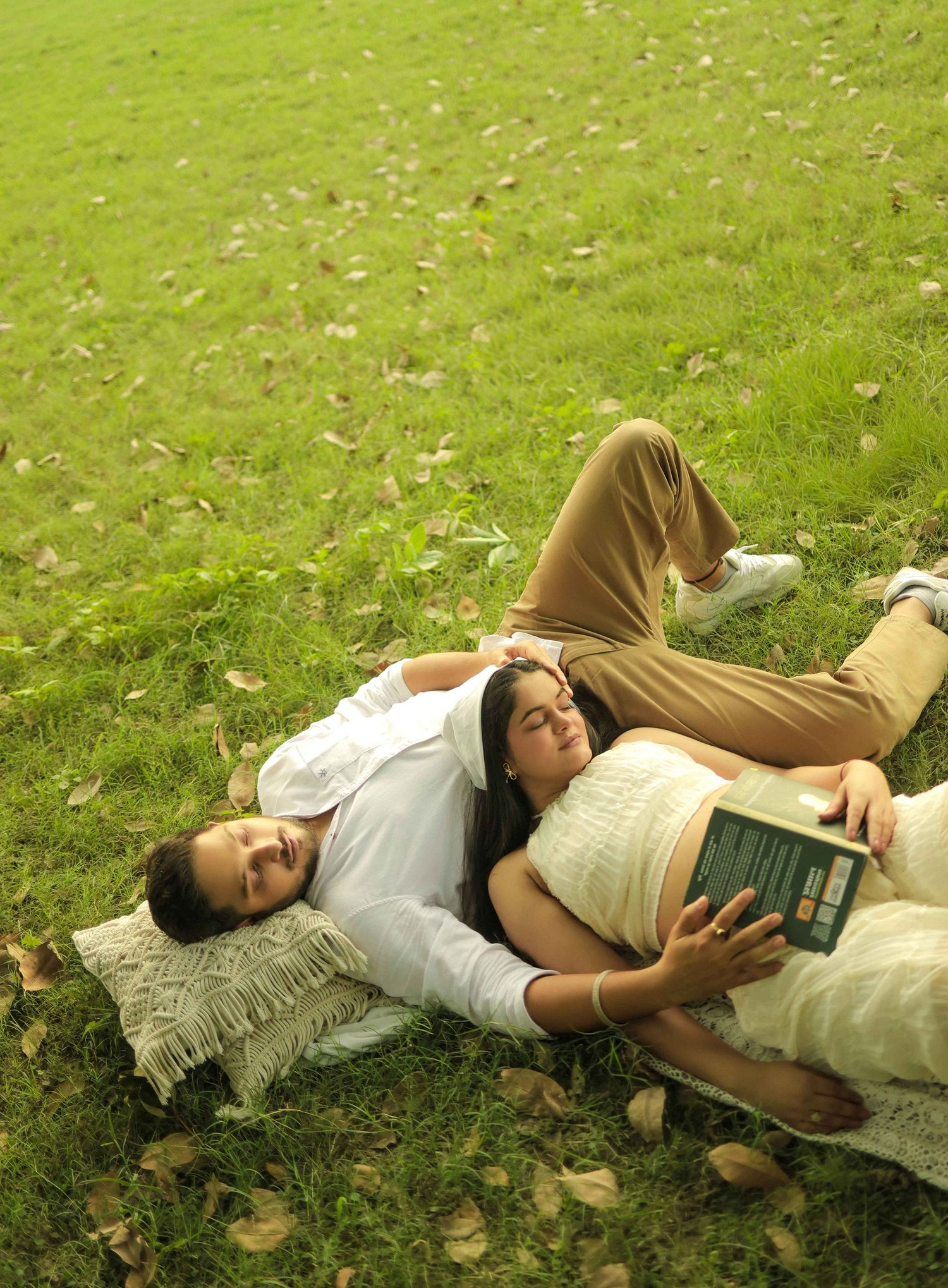 Couple relaxing on green grass, woman resting head on man's chest while holding a book outdoors.