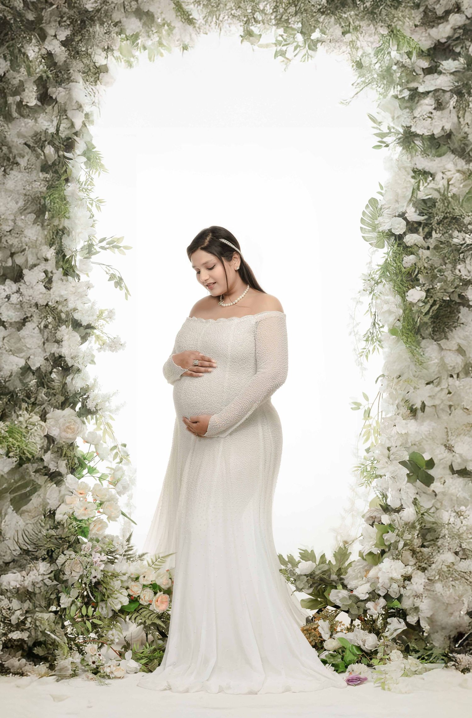 Pregnant woman in white off-shoulder gown standing under floral arch with white flowers and greenery.