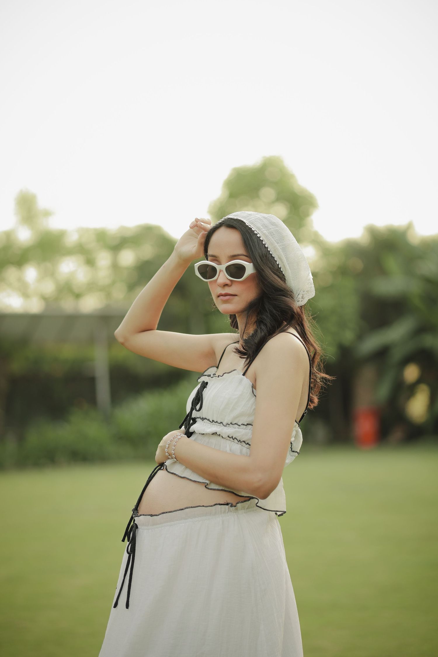 Pregnant woman in white dress and sunglasses posing outdoors on green lawn with trees in background.