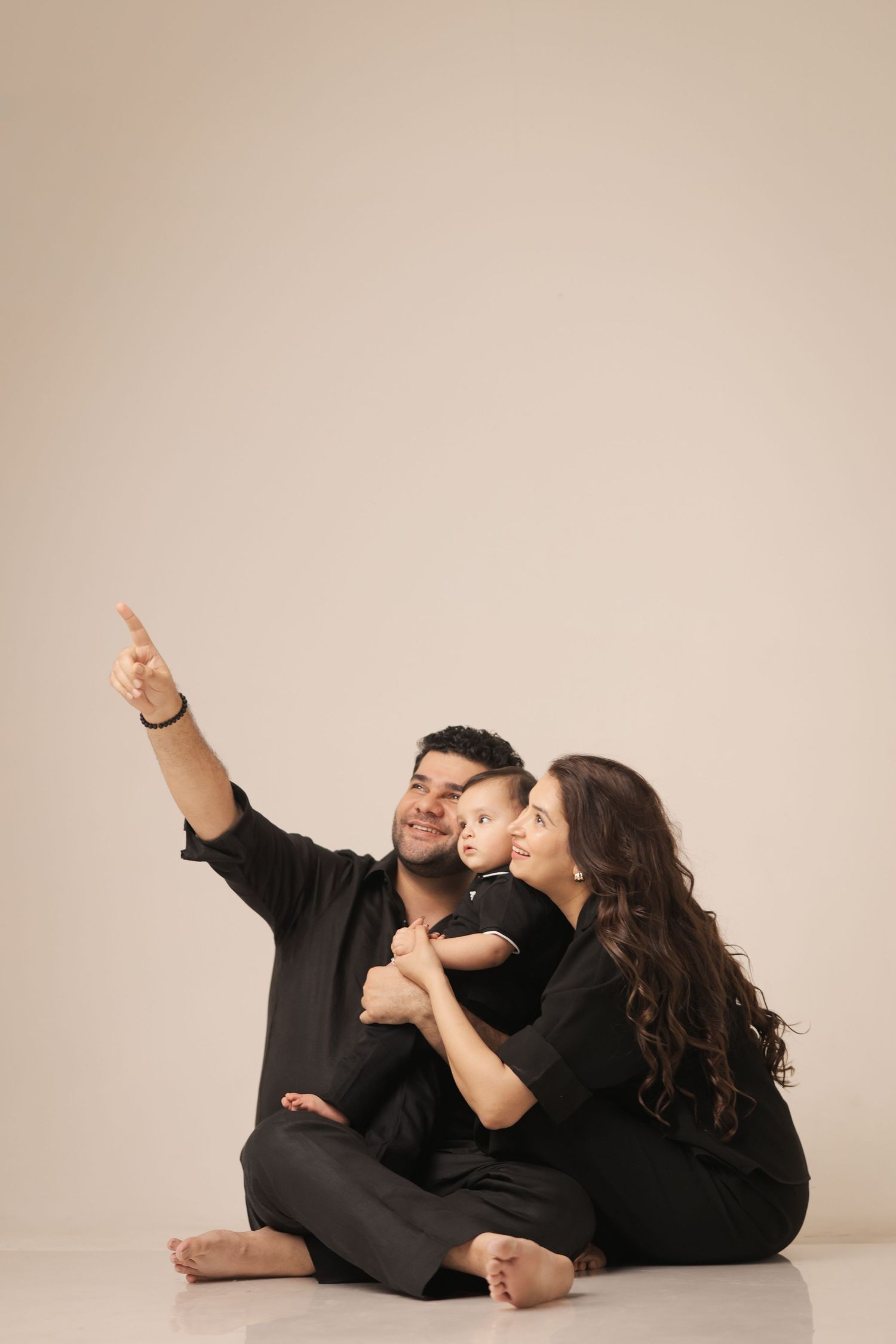 Happy family in black outfits sitting on floor, father pointing up while mother holds smiling baby.