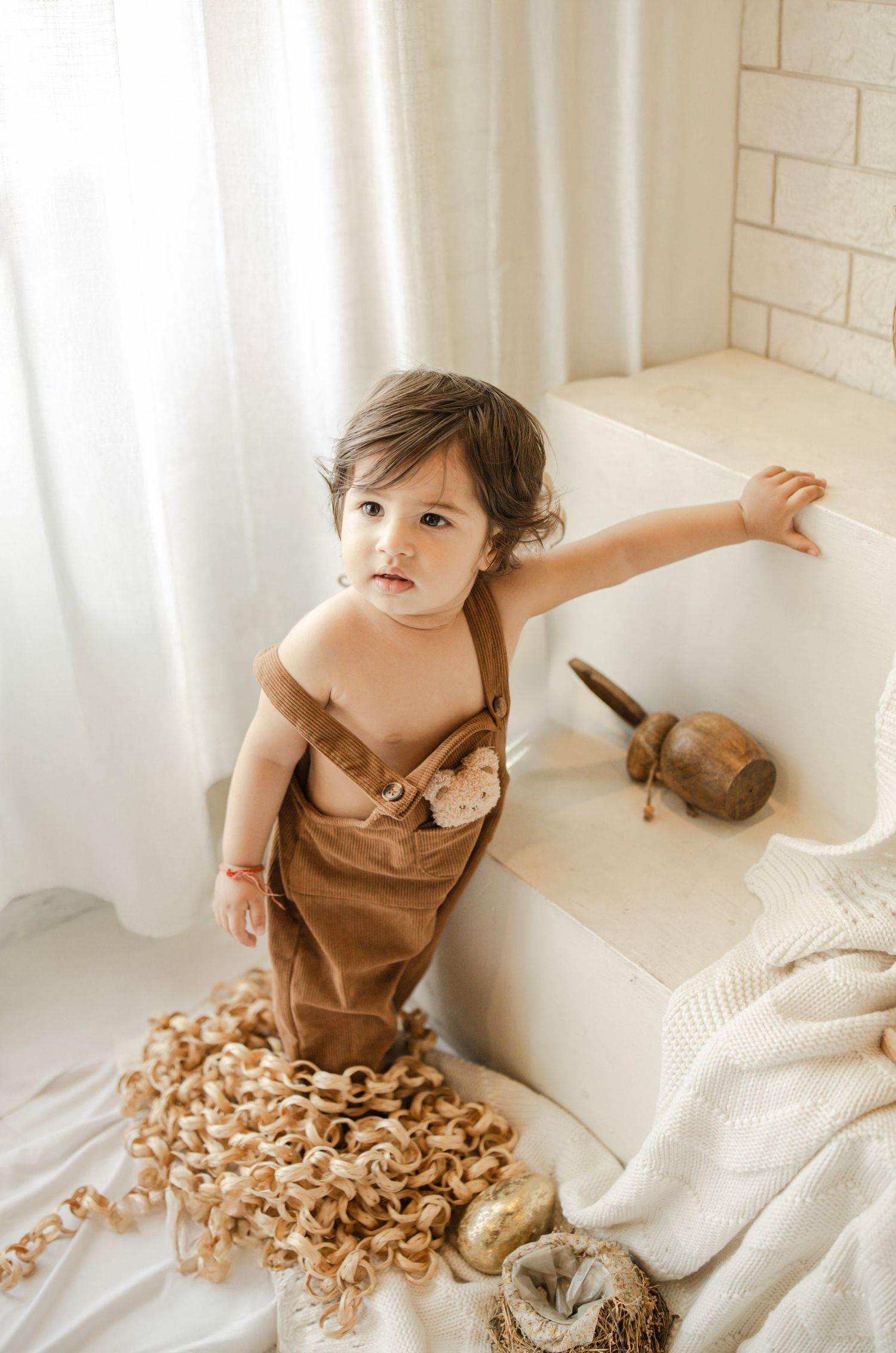 Toddler in brown romper standing by white steps with wooden decor and rattan accents in bright studio.