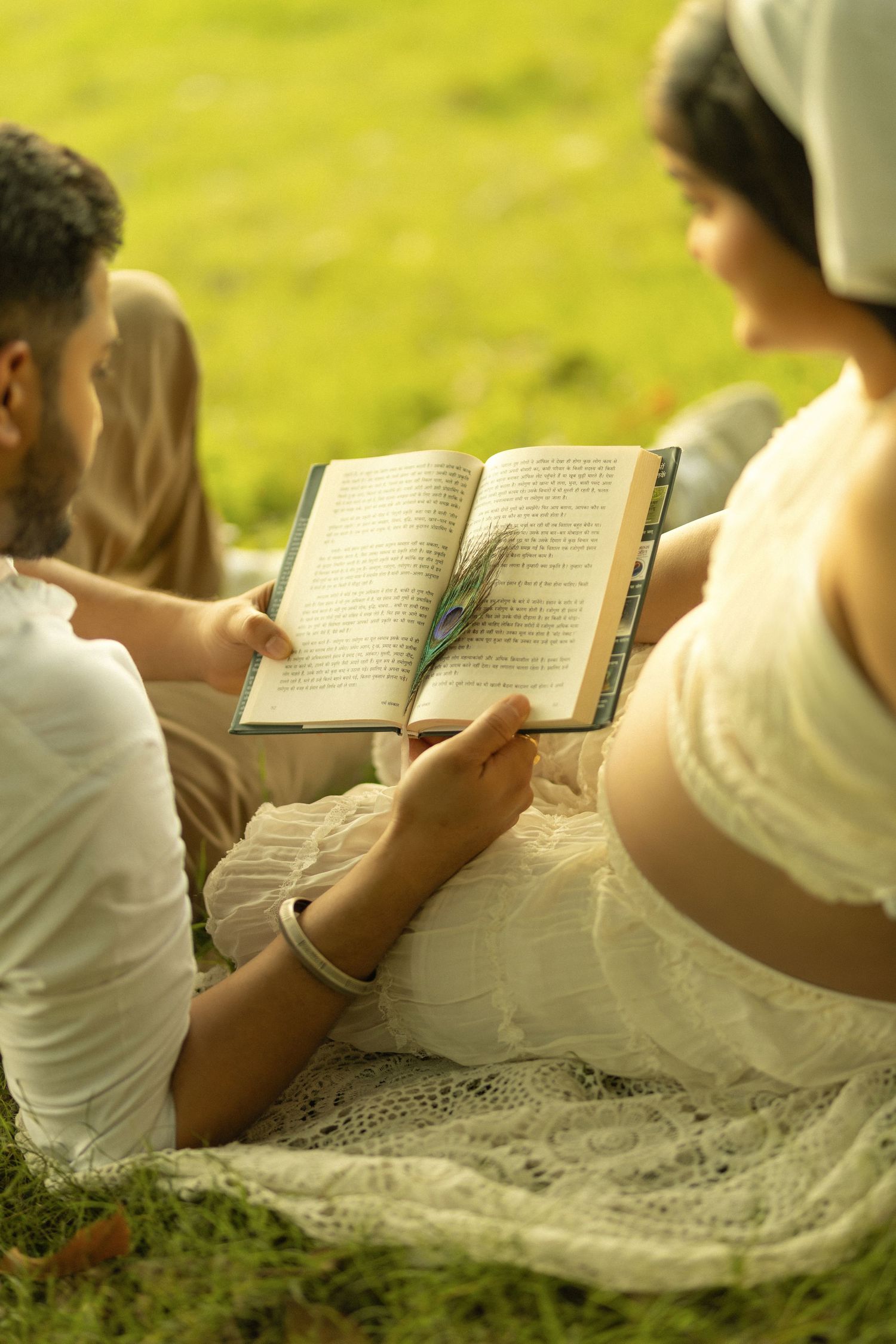 Couple sitting on grass reading an open book together outdoors on a sunny day.