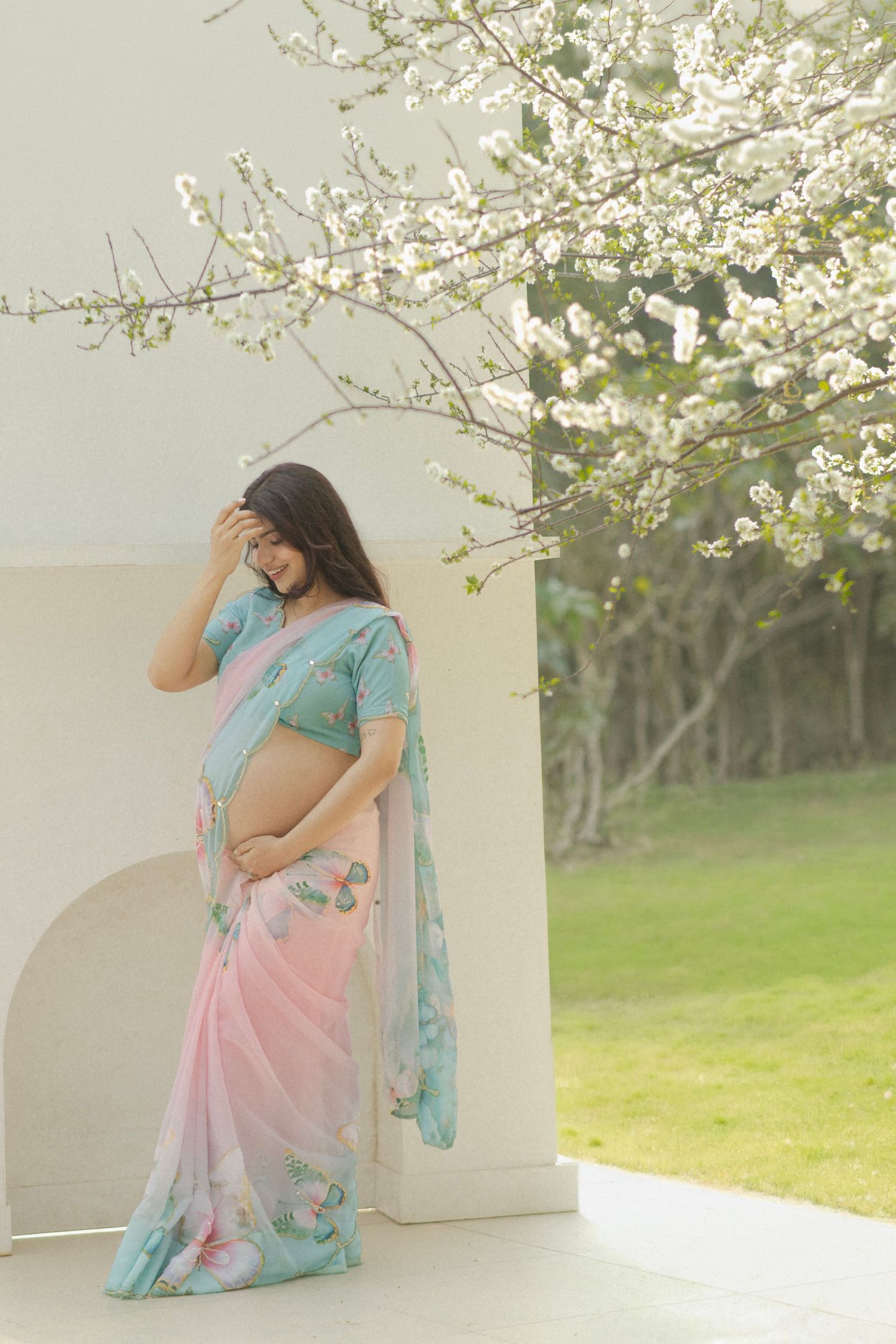 Pregnant woman in pink and teal floral saree standing by white wall with blooming white flowers.