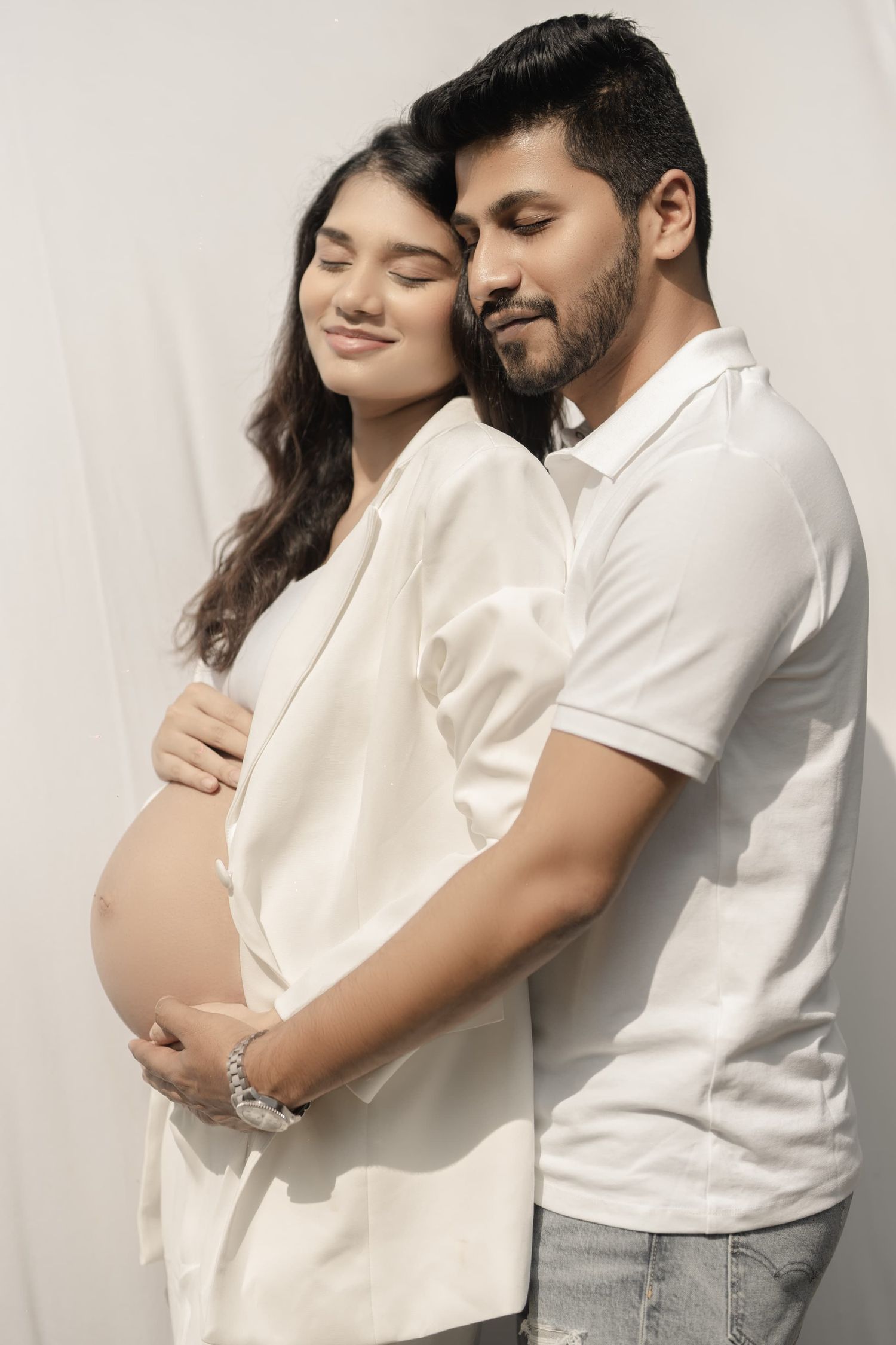 Intimate pregnancy photoshoot featuring couple in white against light background.