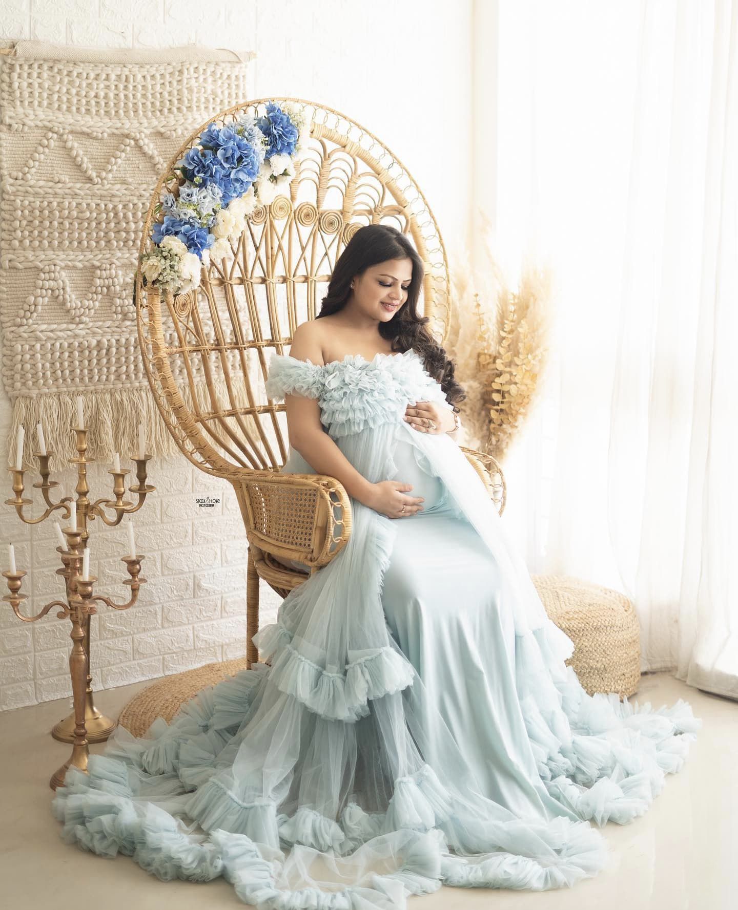 Maternity portrait in a bright indoor setting with a peacock chair and light blue flowing dress.