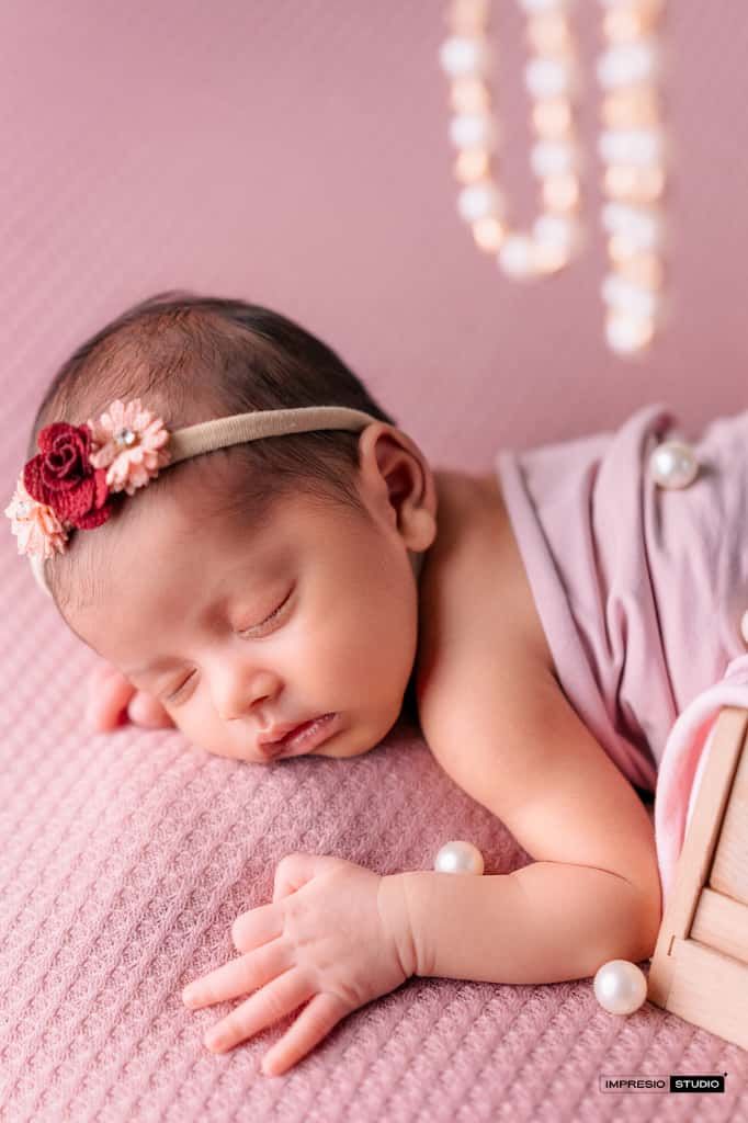 Sleeping newborn baby girl wearing a floral headband on a pink textured blanket in a studio photography setting.