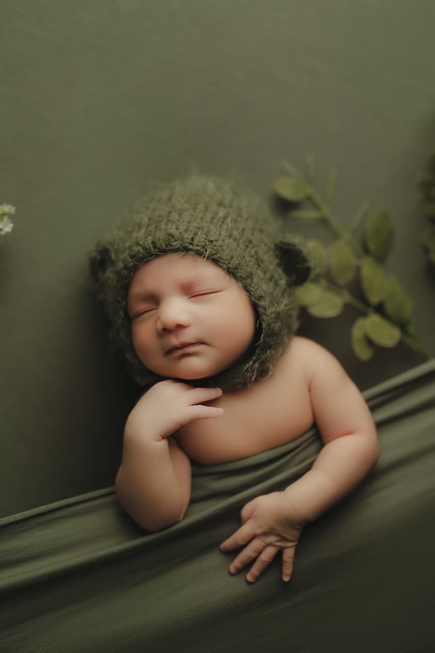 Sleeping newborn baby wearing a green knit bear bonnet hat, wrapped in green fabric against a green backdrop.