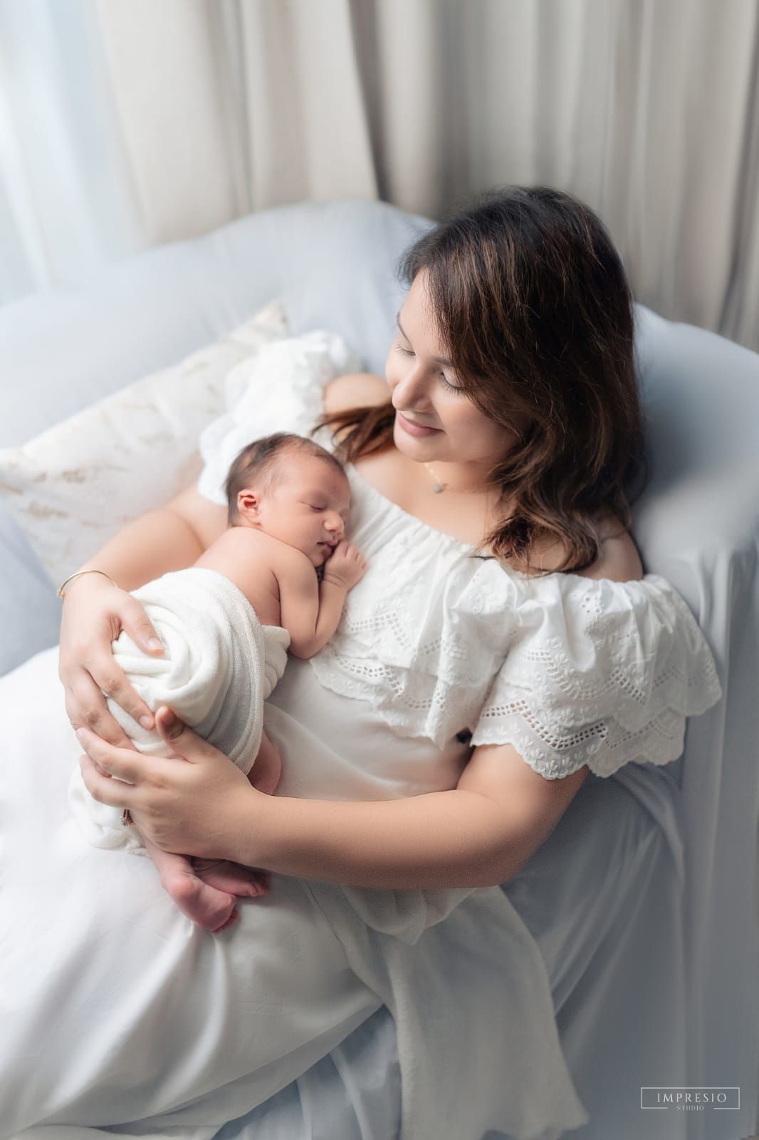 A mother in a white dress tenderly holds her swaddled newborn baby while reclining on a white bed.