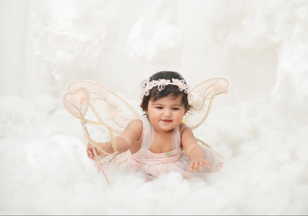 Baby girl in pink tutu dress with fairy wings and floral headband poses among white clouds with a wand.