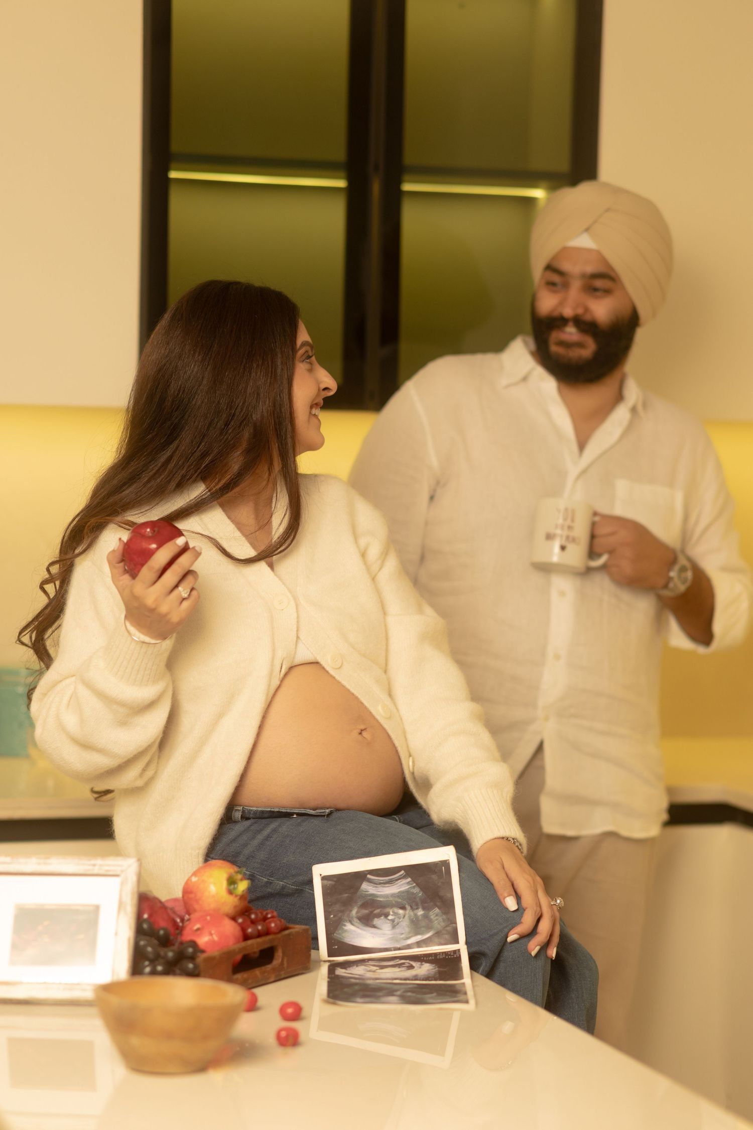 A couple shares a joyful moment in their kitchen while looking at ultrasound photos and fresh fruit on the counter.