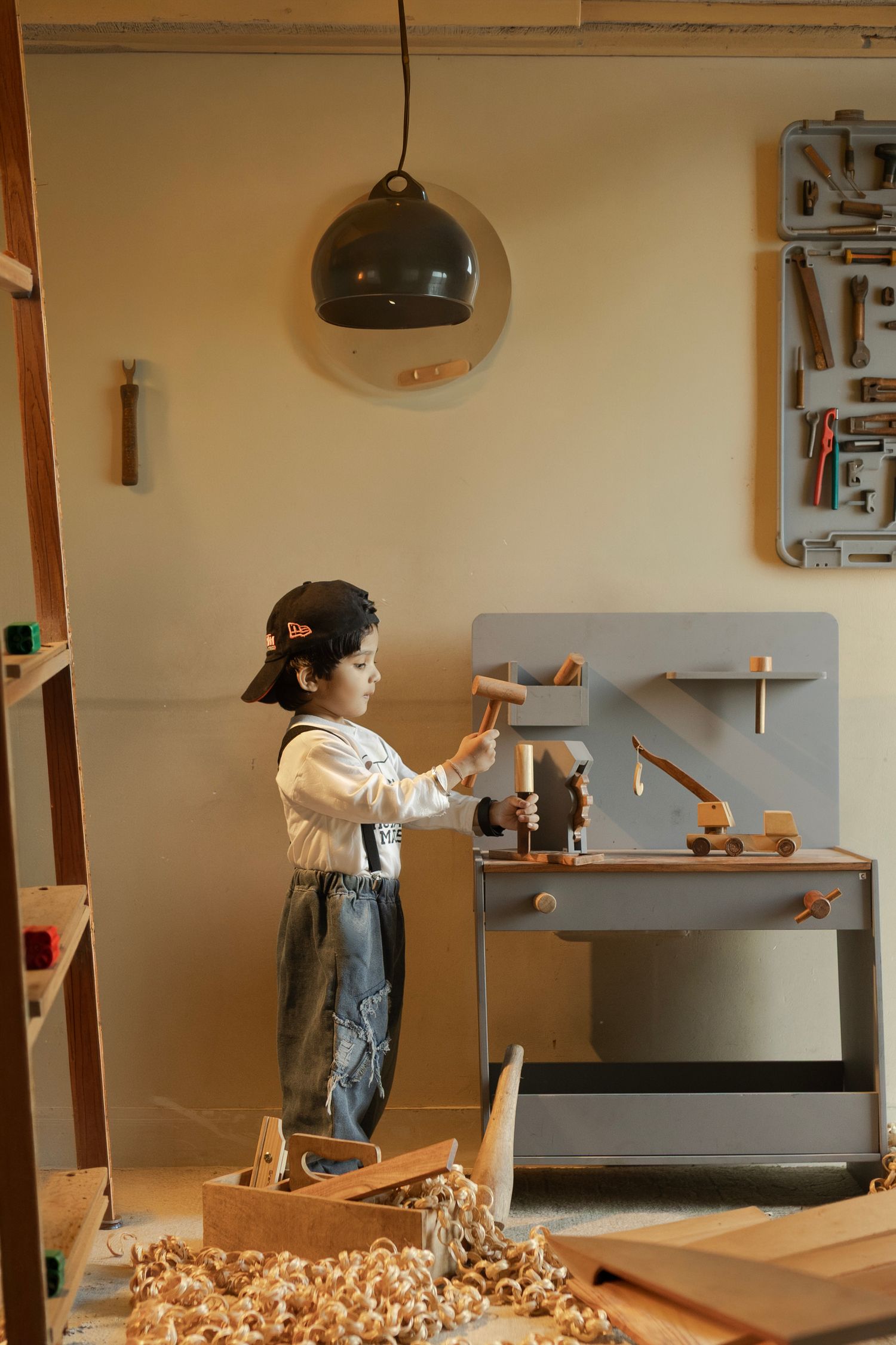 Young child playing with wooden toy workbench surrounded by wood shavings in a cozy carpentry workshop.
