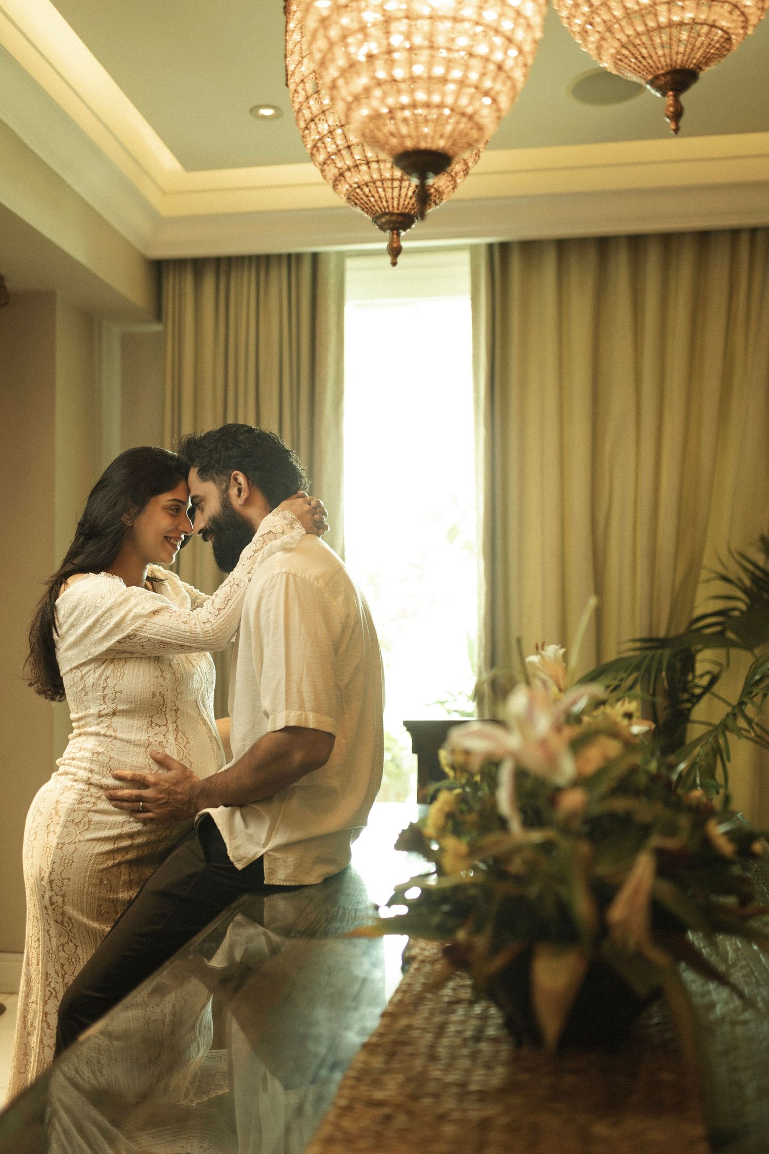 Couple embraces in an elegant room with crystal chandeliers and sunlit windows as potted plants decorate the space for maternity photoshoot.