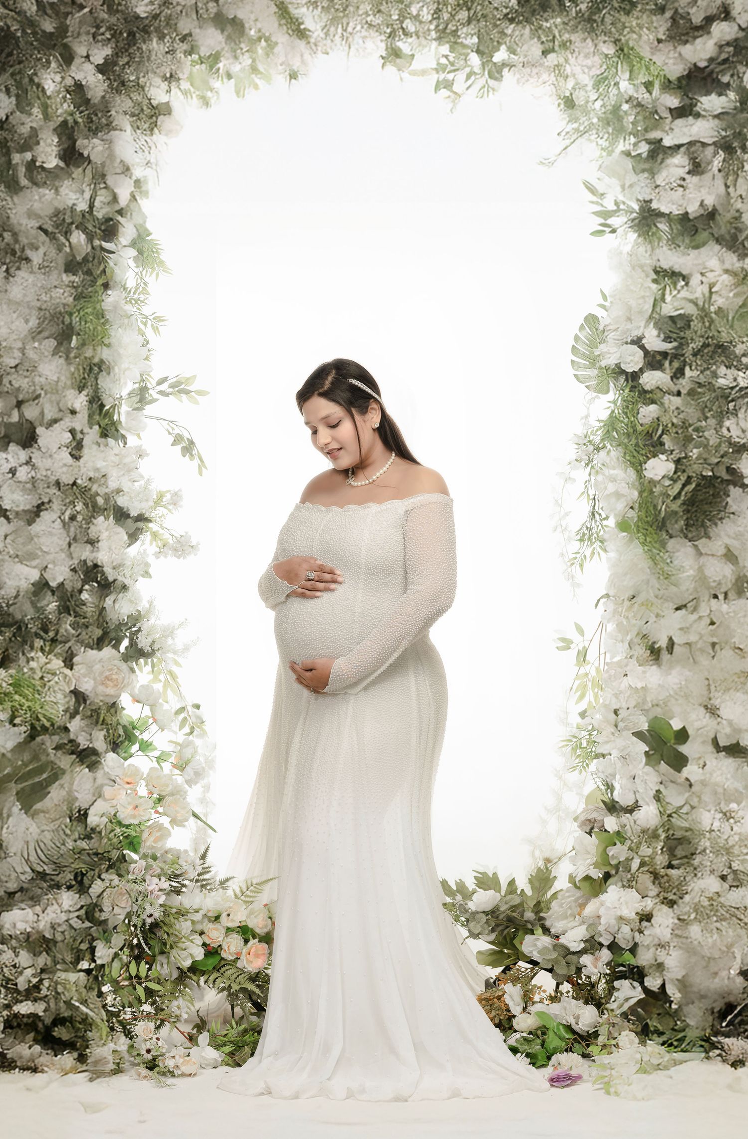 Elegant maternity photography series with flowing white gown against a backdrop of delicate floral arches and greenery.