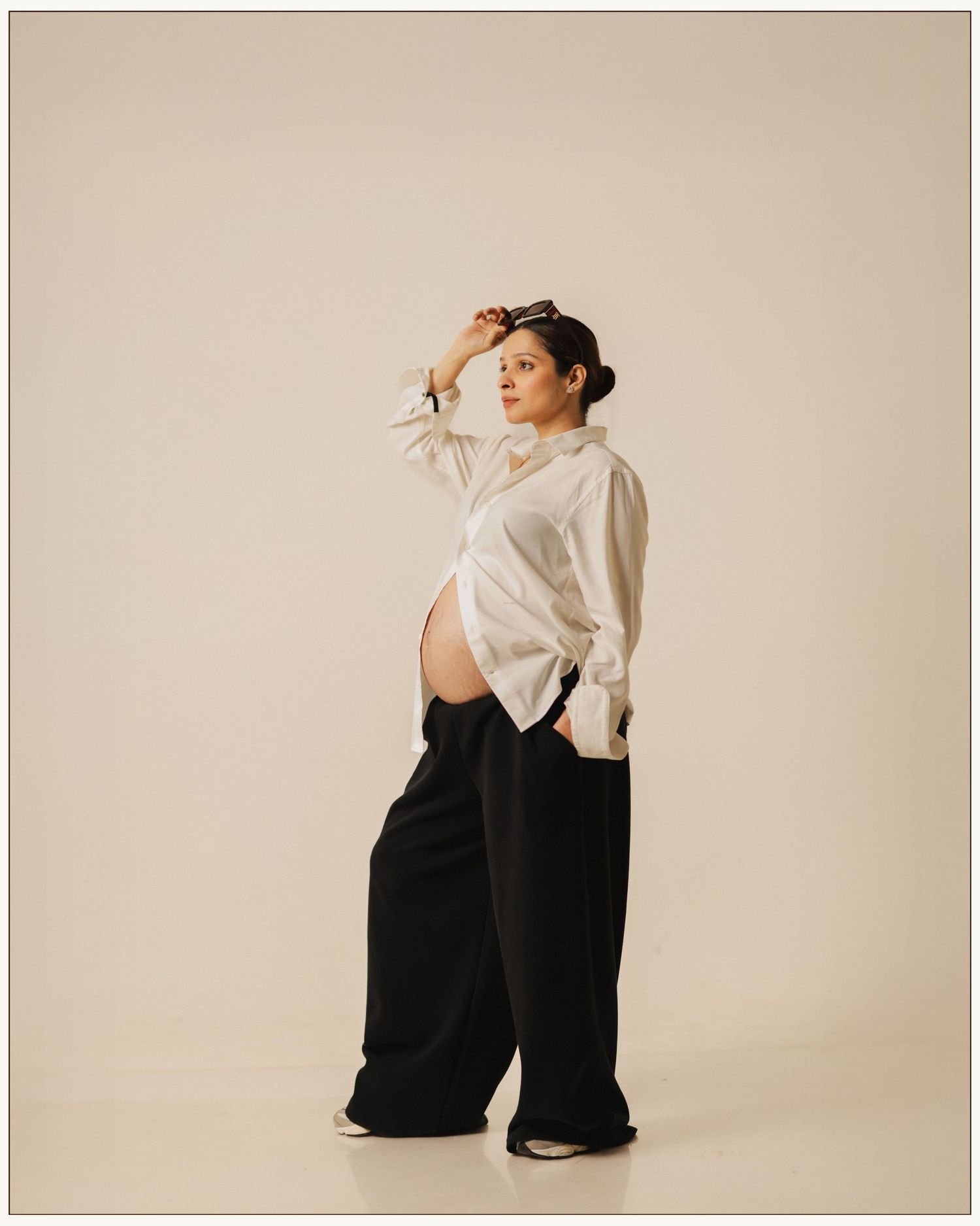 Pregnant woman posing in white shirt and black wide-leg pants against neutral background.