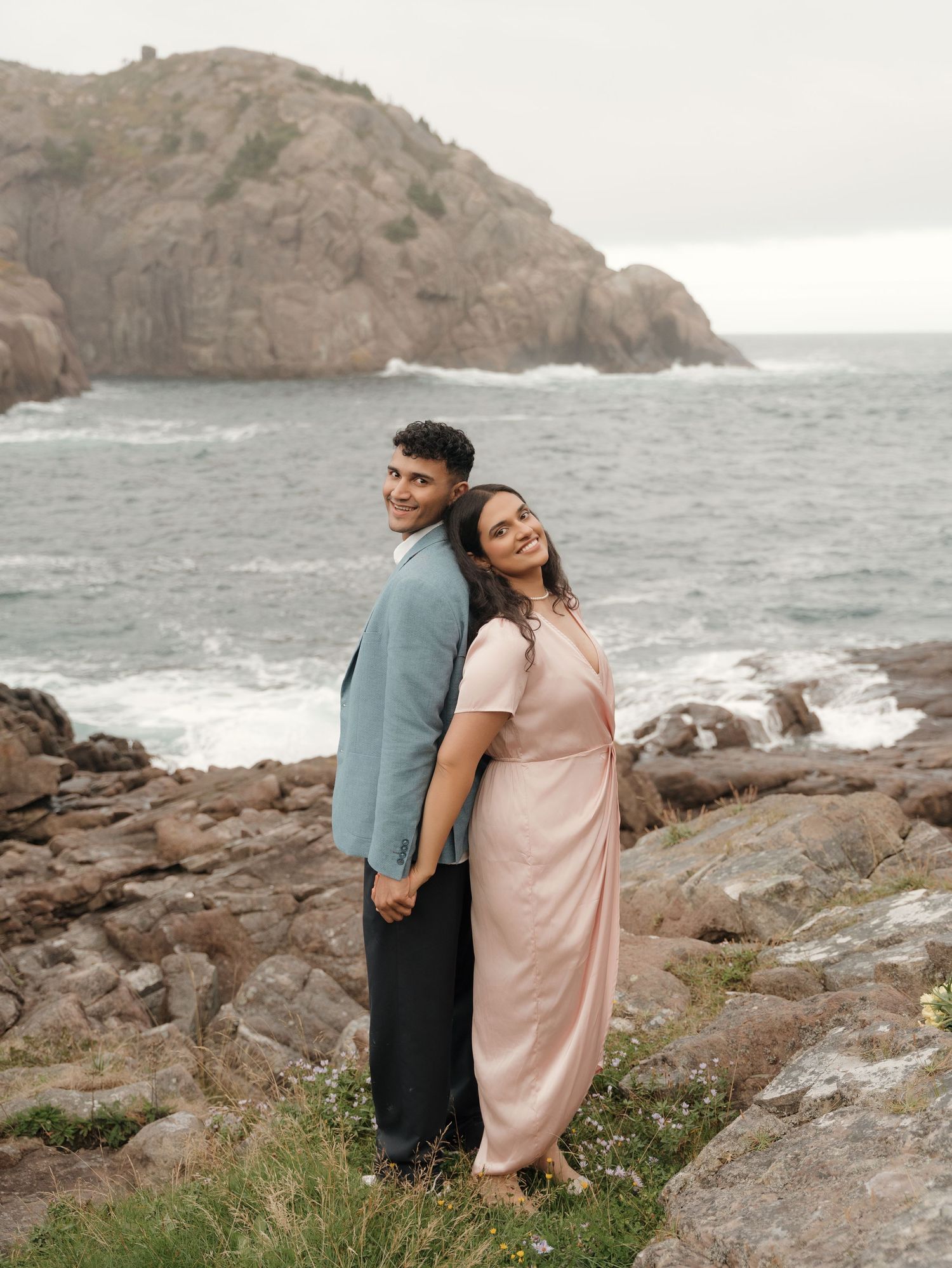 Couple in formal attire sharing romantic moments on rocky coastal cliffs with ocean waves crashing in background.