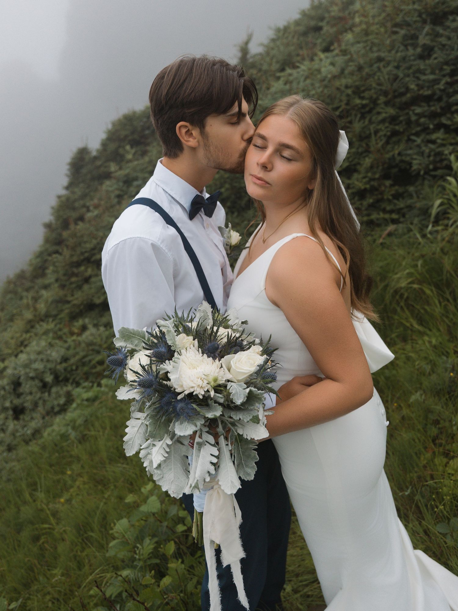 Wedding couple embraces against a scenic coastal backdrop with elegant white floral bouquet.