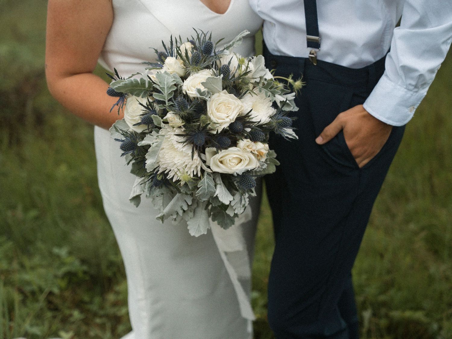 A wedding couple poses with a beautiful white and grey floral bouquet against a natural outdoor background.