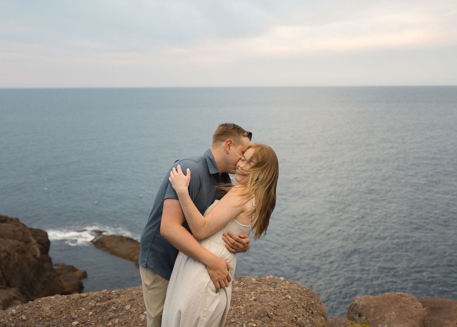 Couple standing on a coastal cliff at sunset sharing a romantic moment overlooking the ocean.
