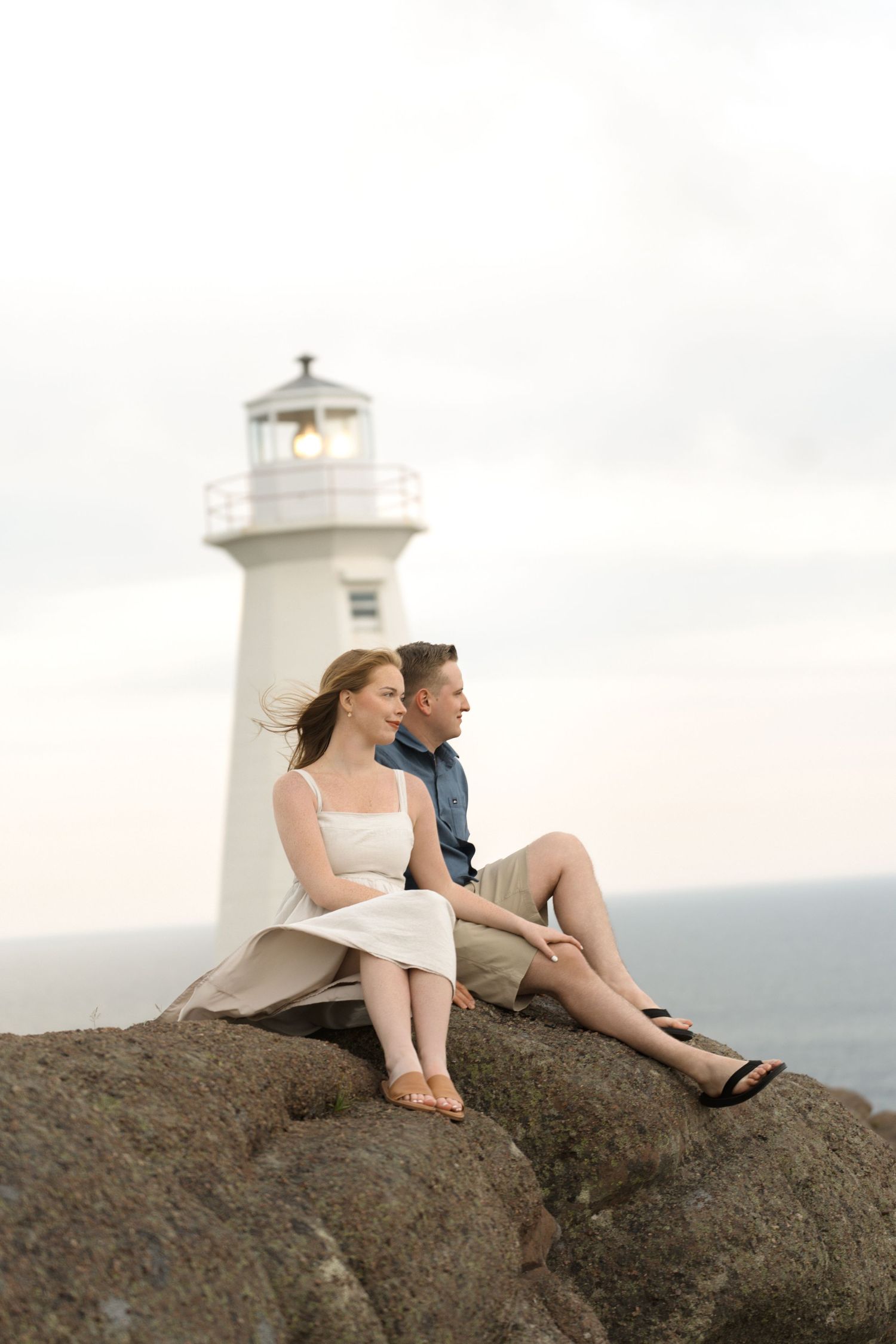 A couple sits together on coastal rocks overlooking the ocean with a white lighthouse in the distance at sunset.