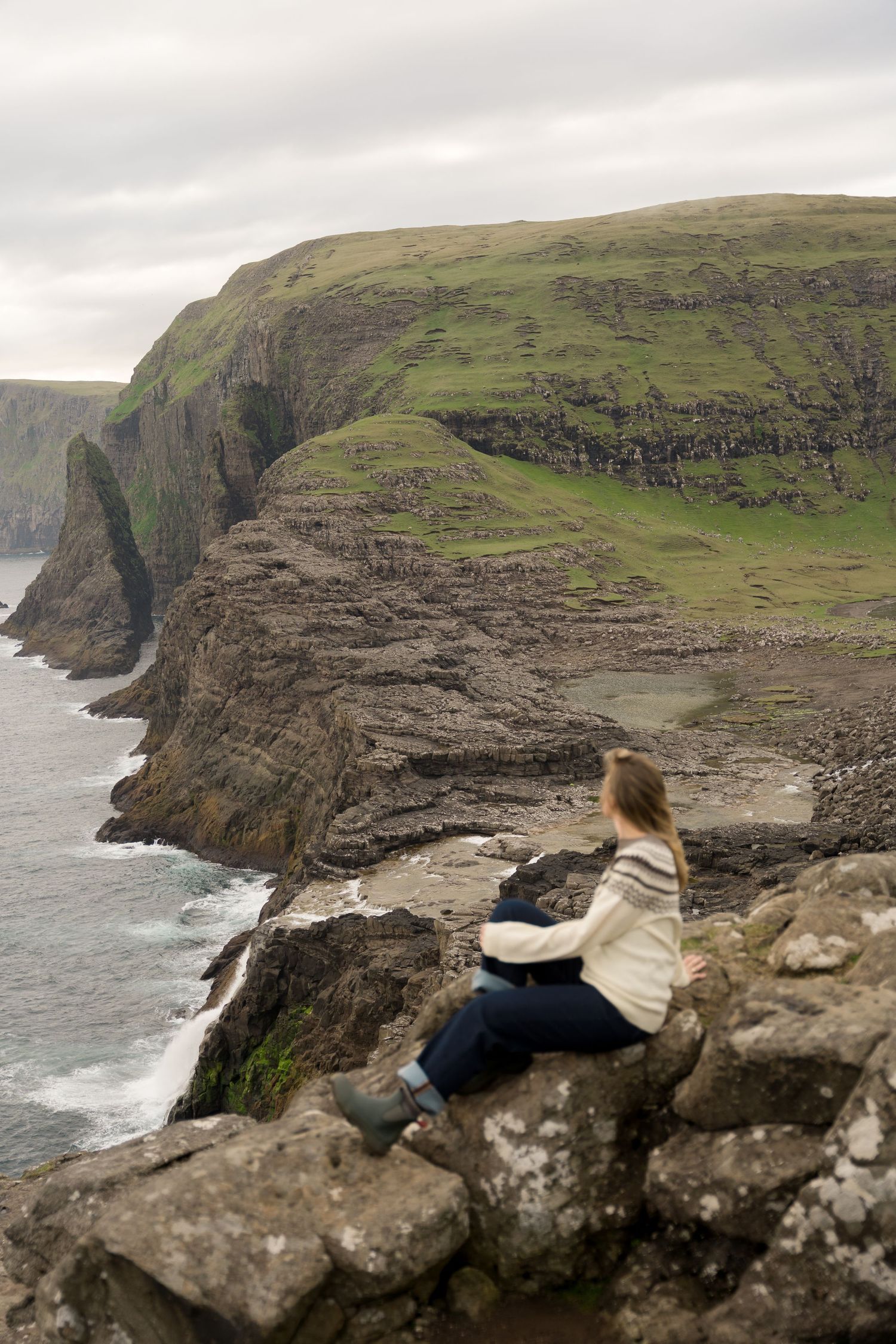 Woman sitting on rocky cliffs overlooking dramatic coastal scenery with green headlands and ocean waves below.