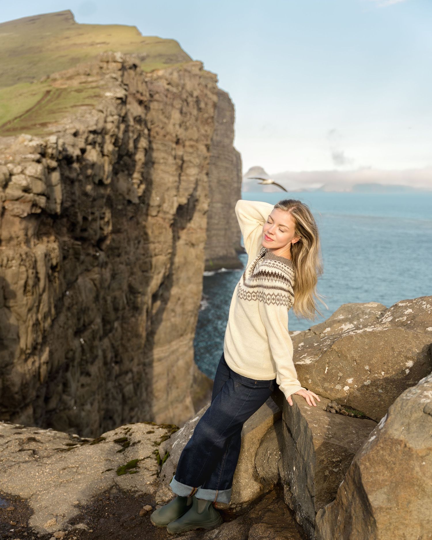 Woman in cream Nordic sweater posing on rocky cliffs overlooking dramatic ocean coastline on a sunny day.