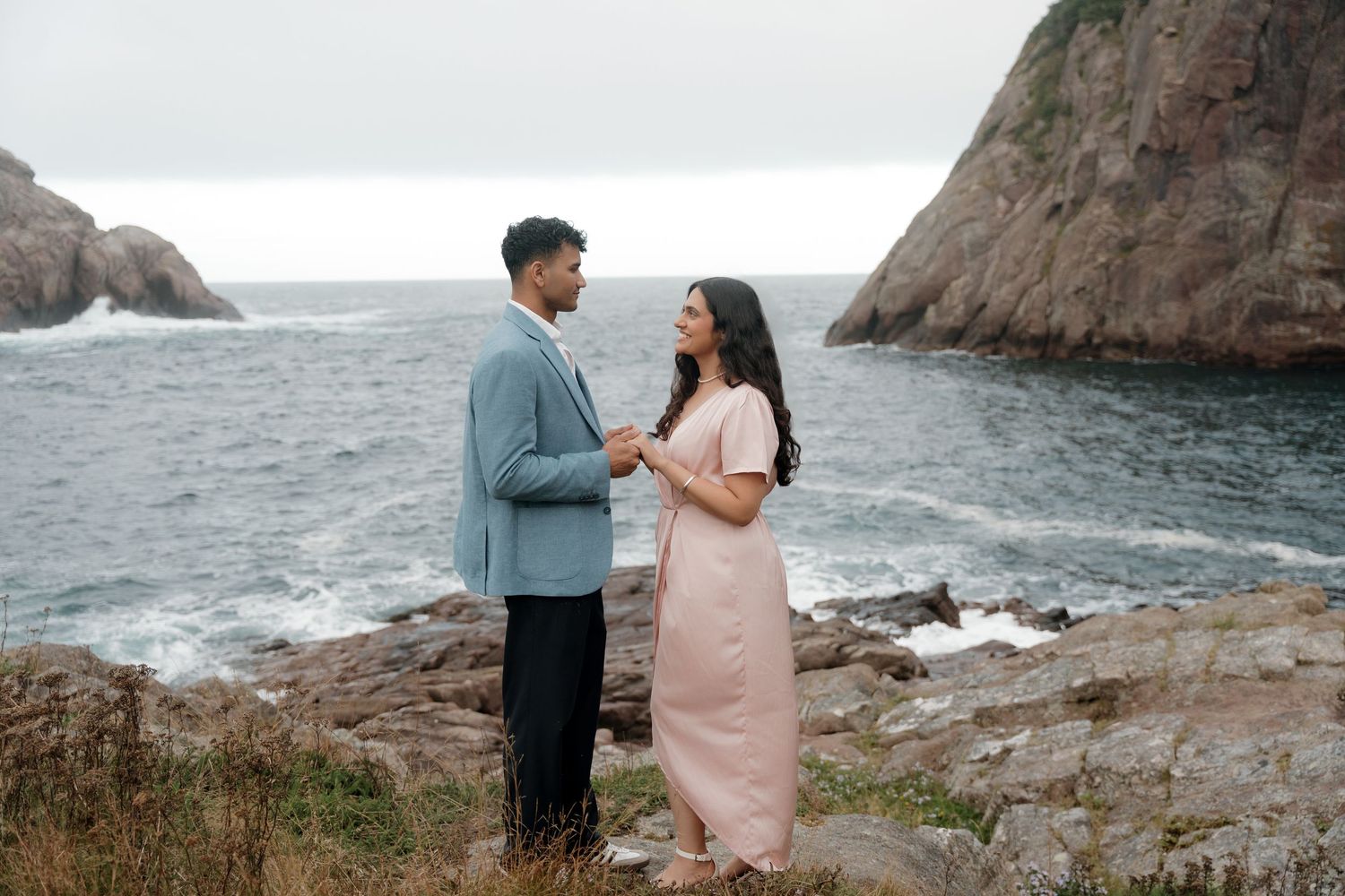 A couple in formal wear shares romantic moments along a rugged coastal cliff with dramatic ocean views in the background.