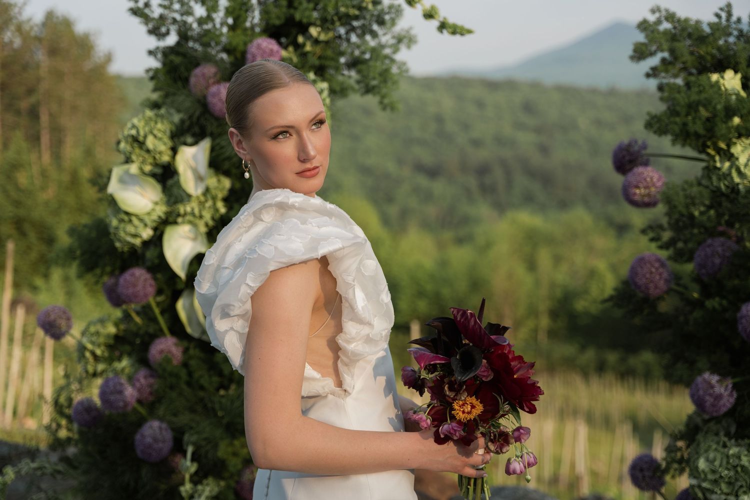Elegant bride in lace dress holds burgundy bouquet with mountains and trees as backdrop.