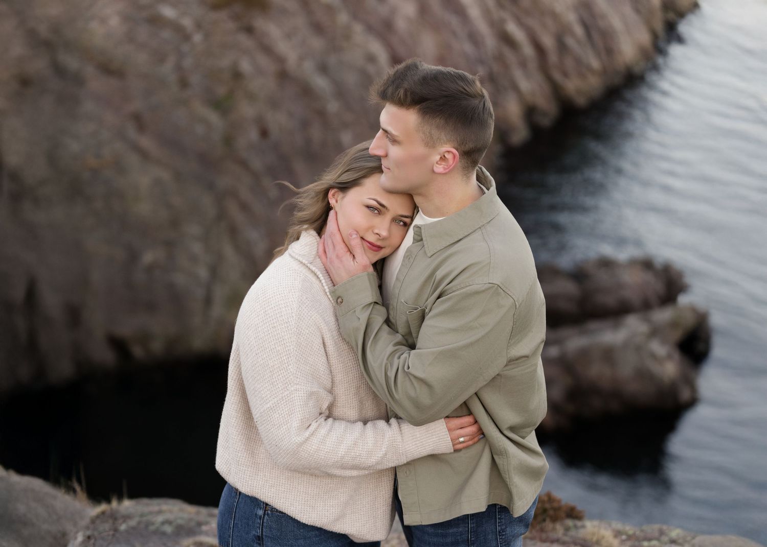 A romantic coastal portrait showing an intimate embrace against rocky cliffs.