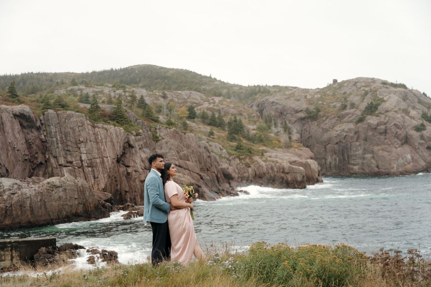 Engaged couple embraces in front of dramatic coastal cliffs and crashing ocean waves at sunset.
