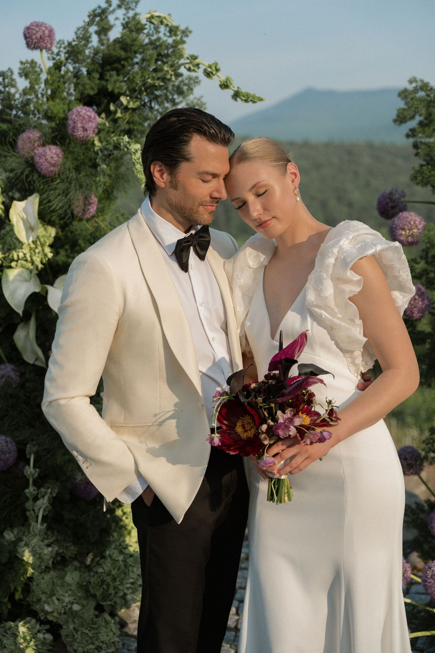 A romantic outdoor wedding scene with a couple in elegant white attire posing together against a mountain backdrop.
