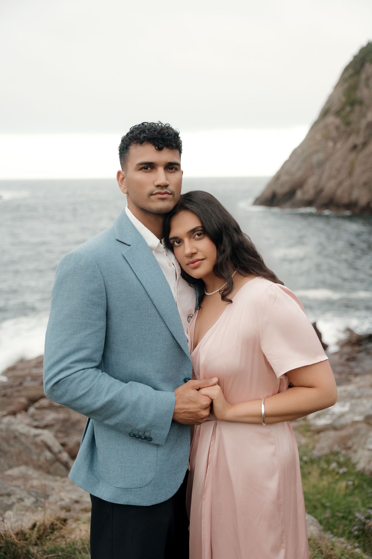 A couple poses together by coastal cliffs, wearing formal attire in soft blue and pink tones against an ocean backdrop.