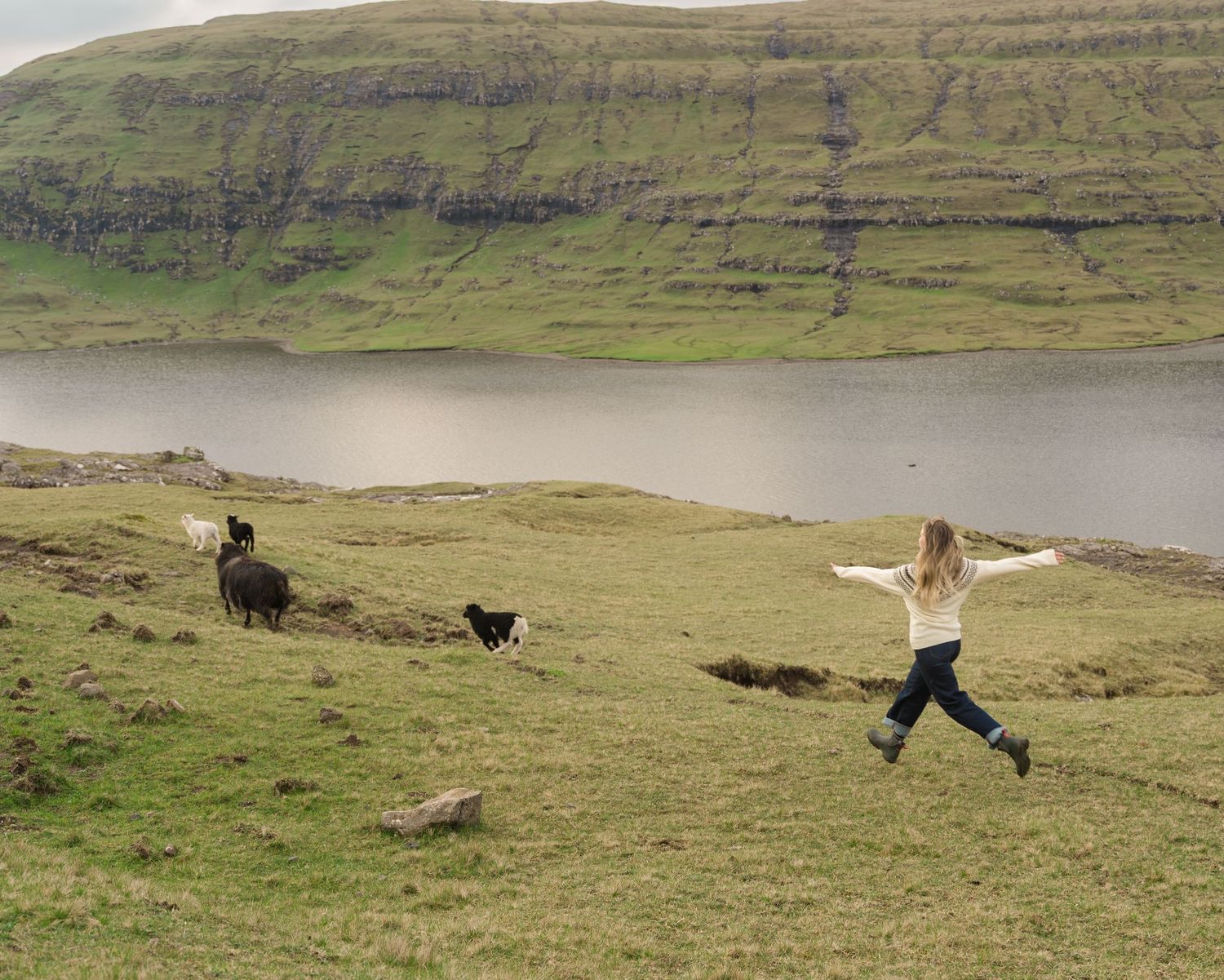 Woman jumping joyfully near sheep on green hillside by a scenic lake with dramatic cliffs in background.