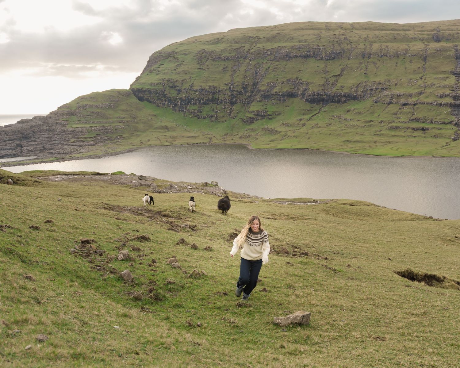 Woman running down a lush green hillside with sheep grazing near a scenic fjord and cliff backdrop.