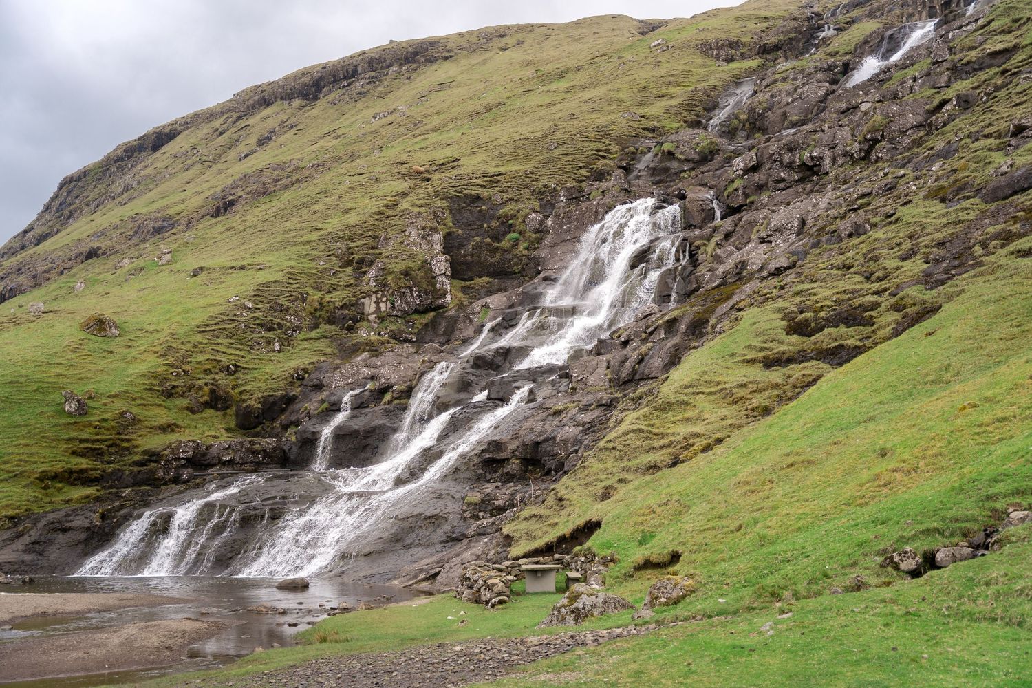 A cascading waterfall flows down a rocky green hillside into a shallow stream in a scenic rural landscape.