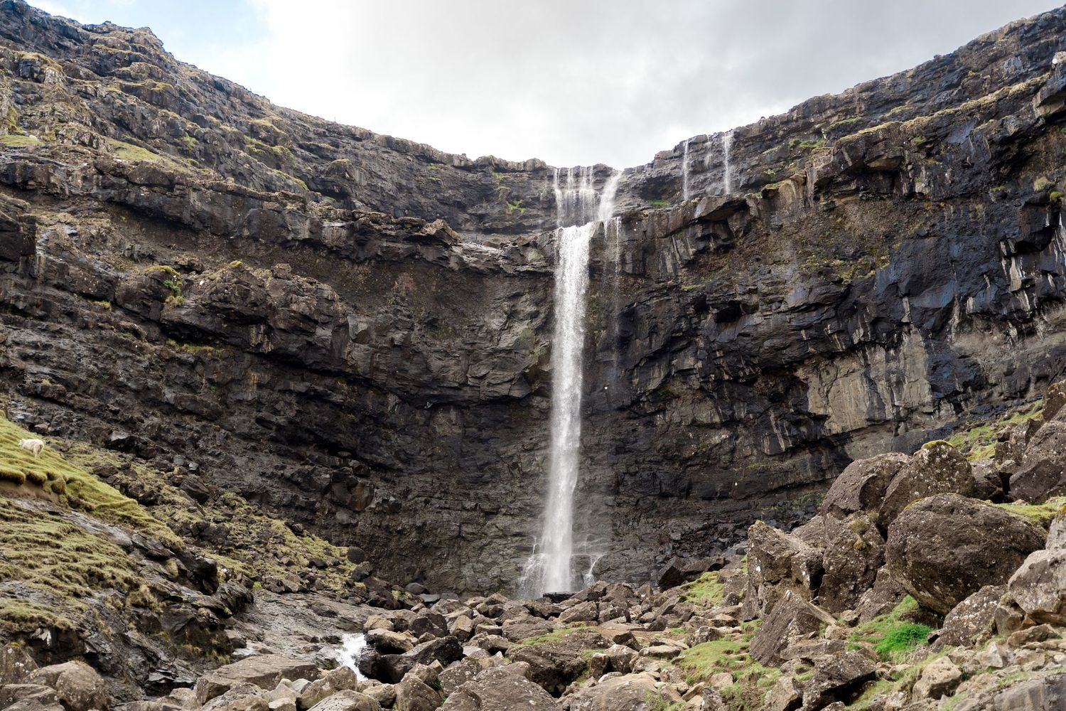 A tall waterfall cascades down a rugged rocky cliff face into a natural amphitheater of dark basalt stone.