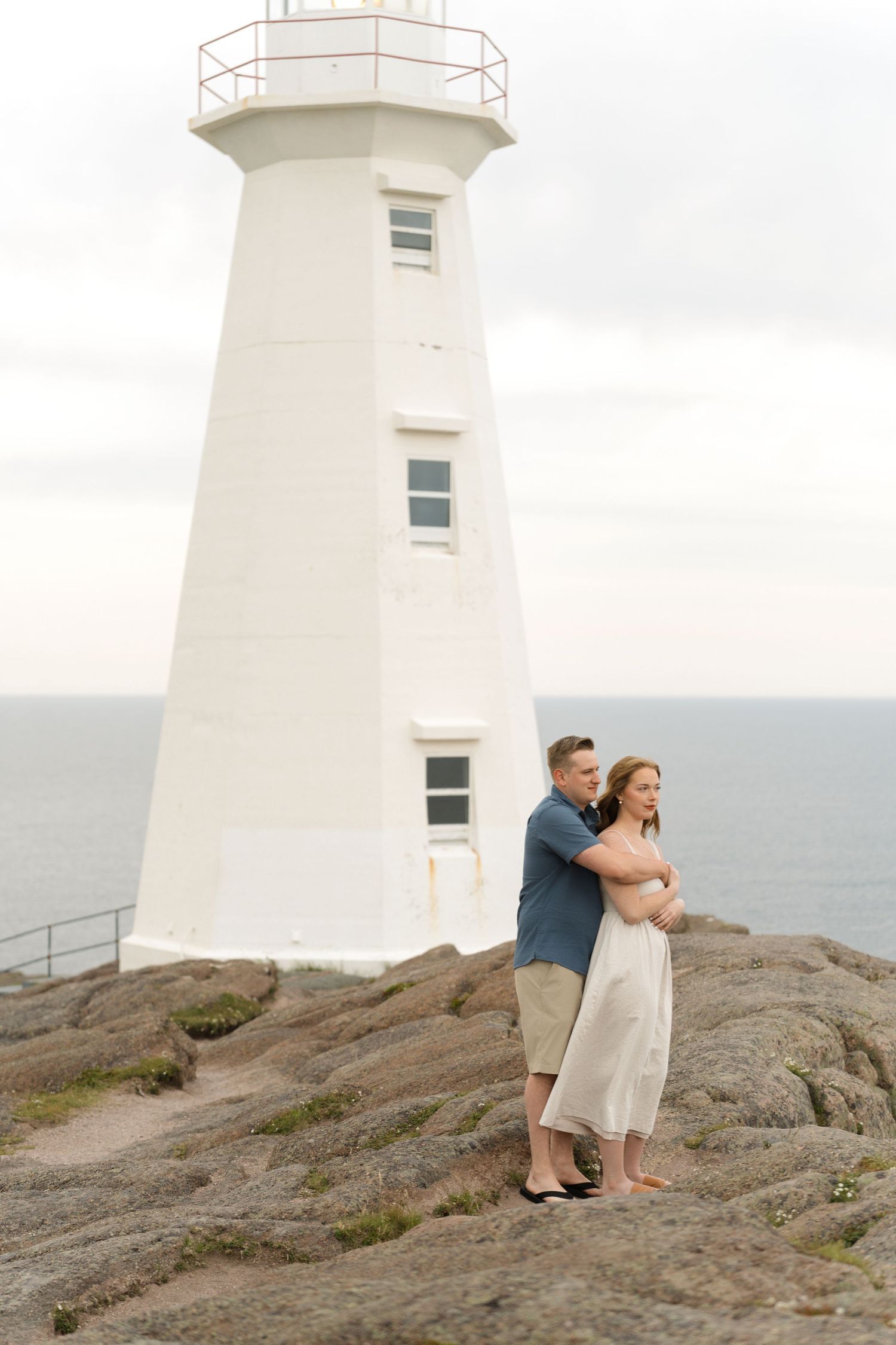 A couple stands together near a coastal lighthouse on rocky terrain against an overcast sky.