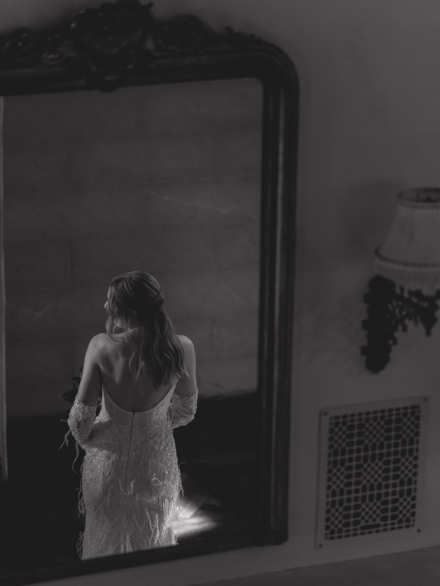 A black and white photograph showing a reflection in an ornate mirror of someone wearing a delicate lace dress.