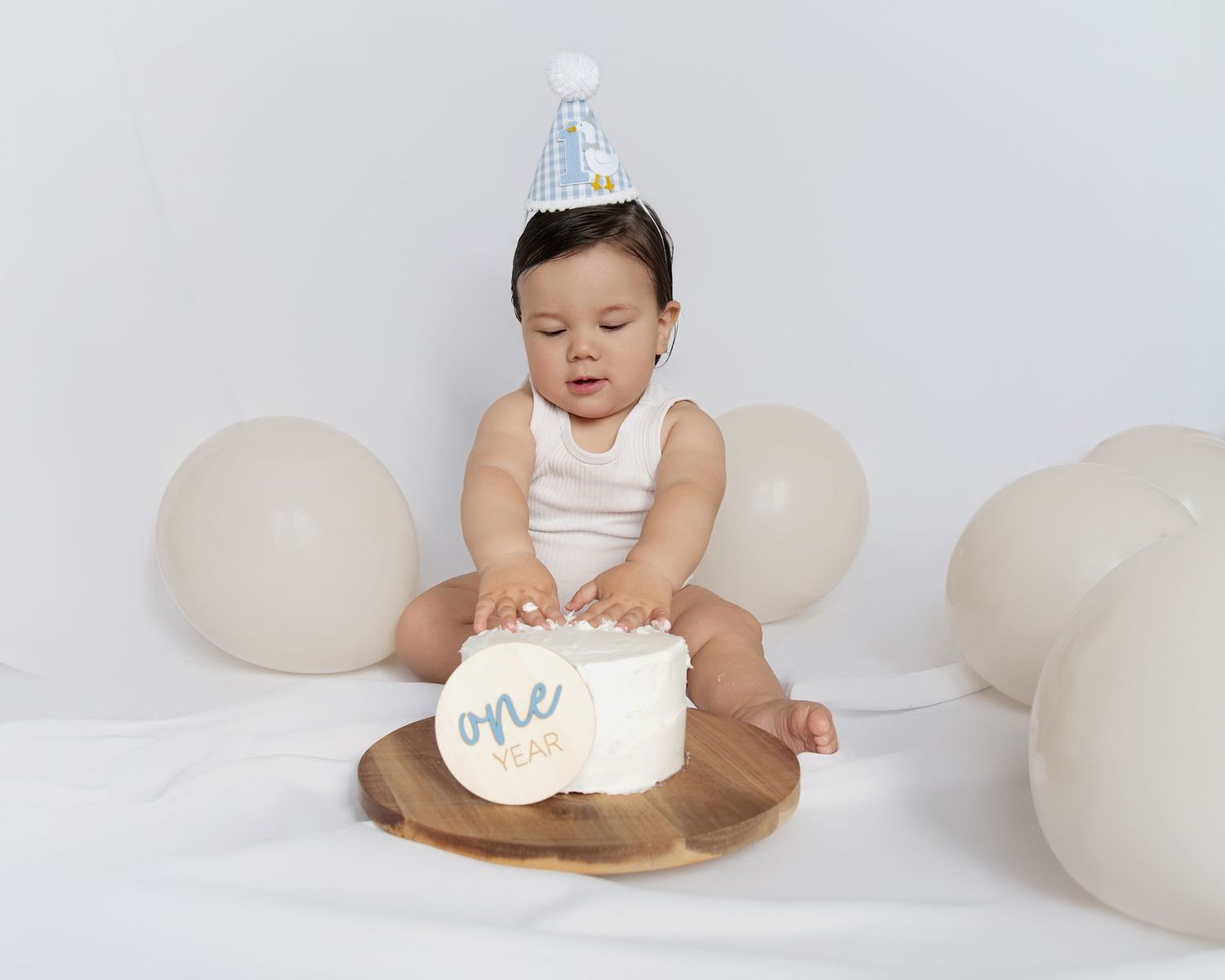 Baby in white outfit sits on beige background with birthday cake and balloons for first birthday photoshoot.